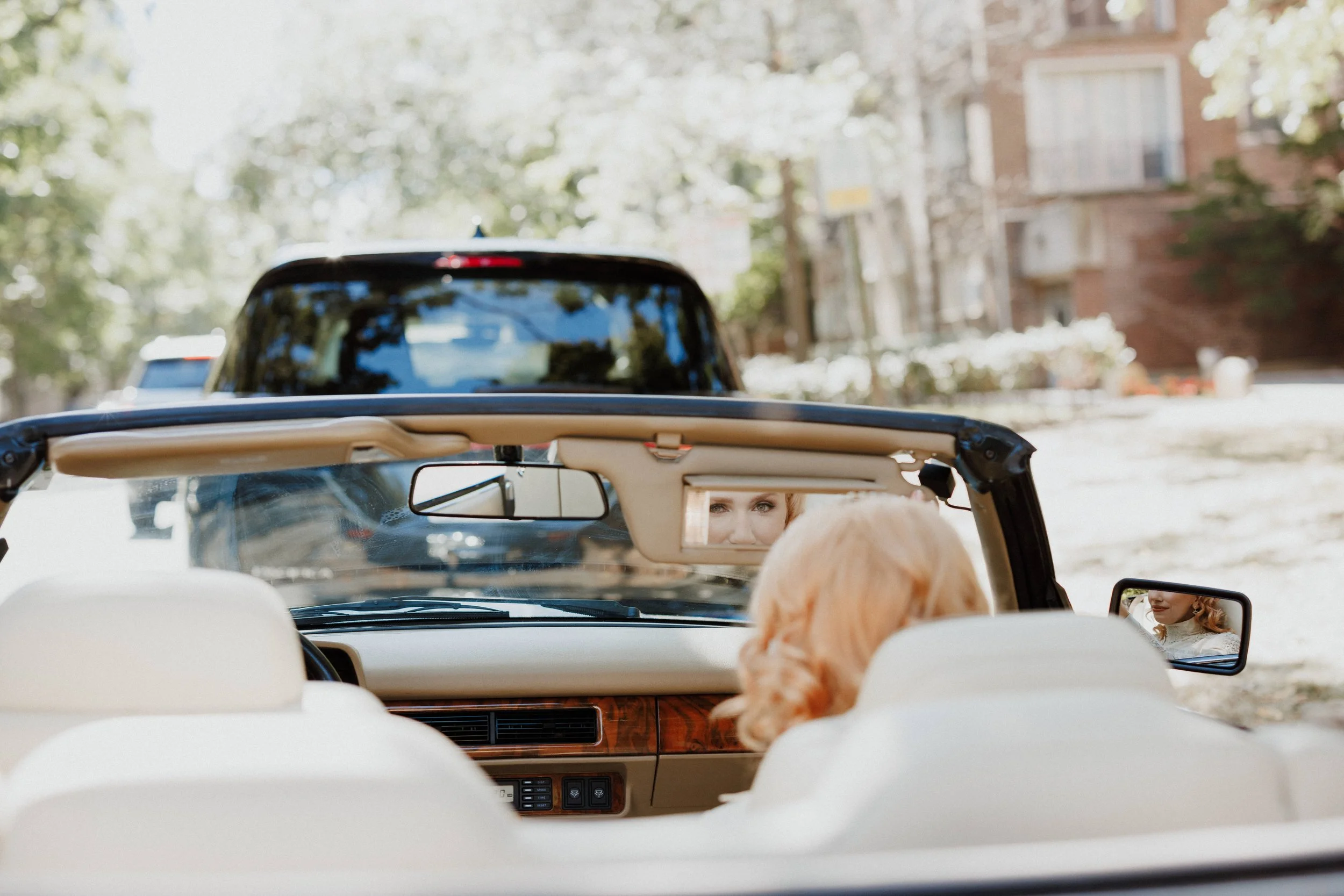 View from inside a convertible car showing a woman with blonde hair in the passenger seat and another woman with red hair in the driver's seat, with a background of trees and buildings.