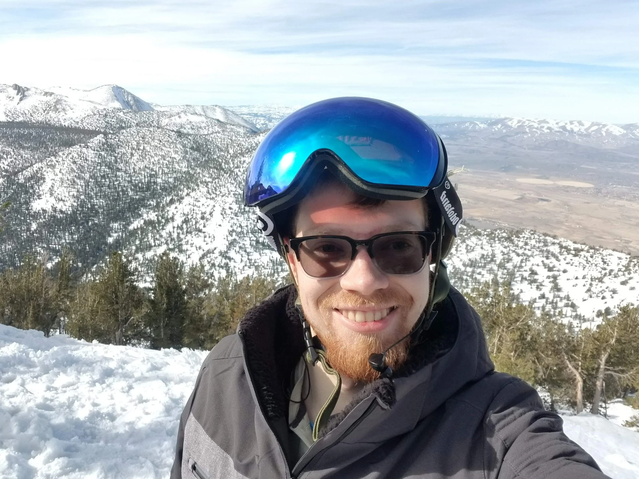 Blaise Jorgensen, candidate for Washoe County Public Administrator,  taking a selfie on a snowy mountain with a helmet and goggles, smiling, with snow-covered mountains and a valley in the background.