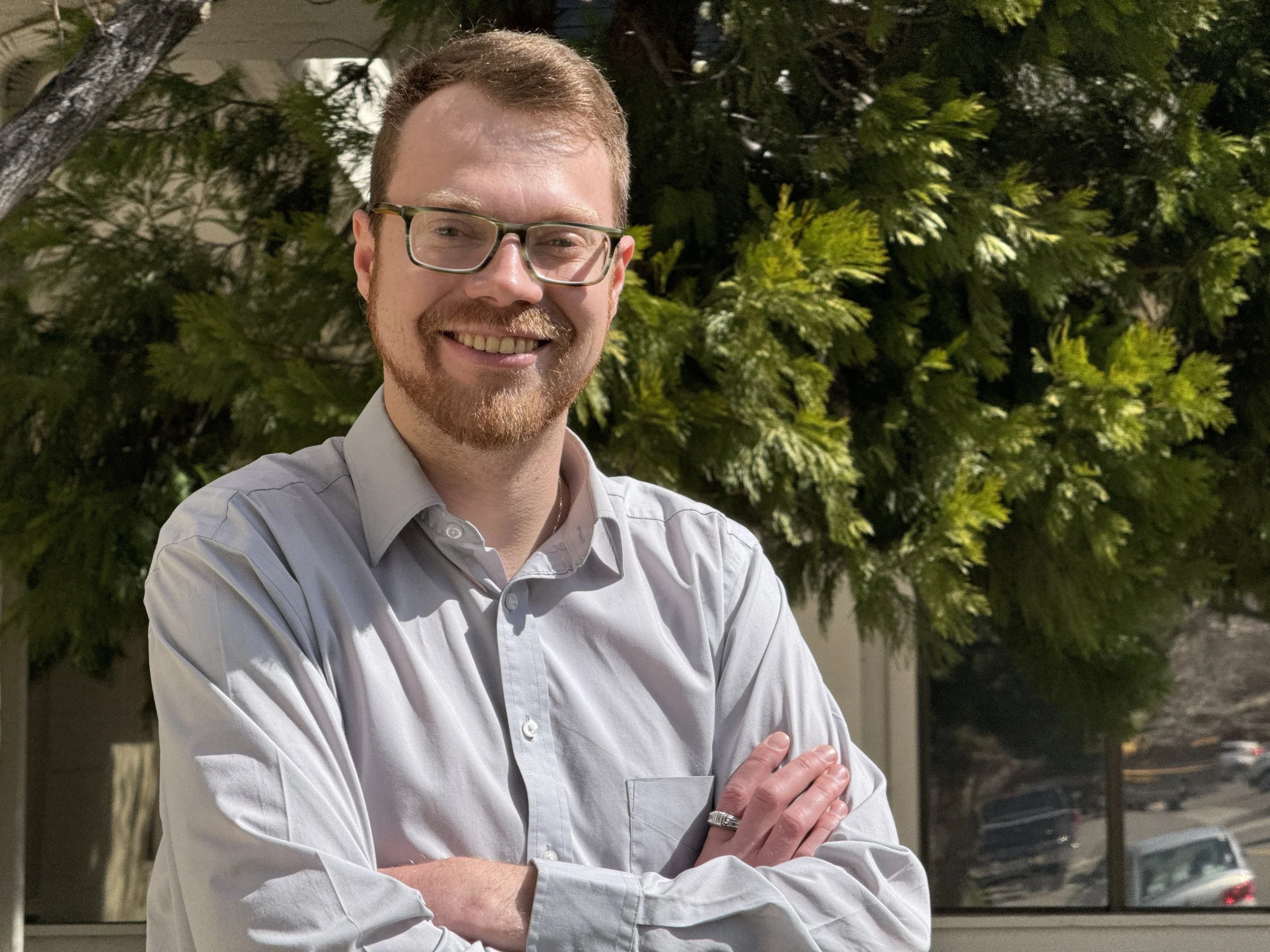 Blaise Jorgensen, candidate for Washoe County Public Administrator, standing outdoors with crossed arms, in front of a tree with green leaves.