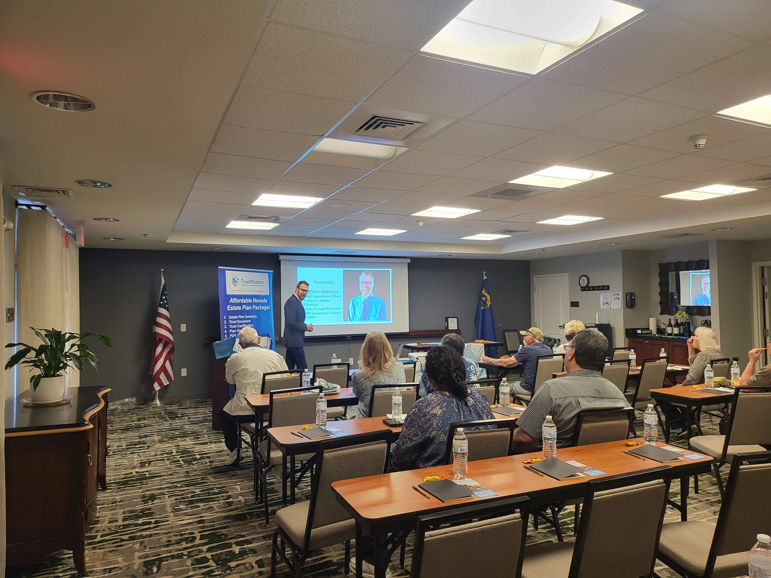 A conference room with people seated at tables, watching a presentation by a speaker at a front podium. The room has an American flag, a TrustMasters banner, and a large screen displaying the presentation. There are water bottles on the tables and a large potted plant near the window.