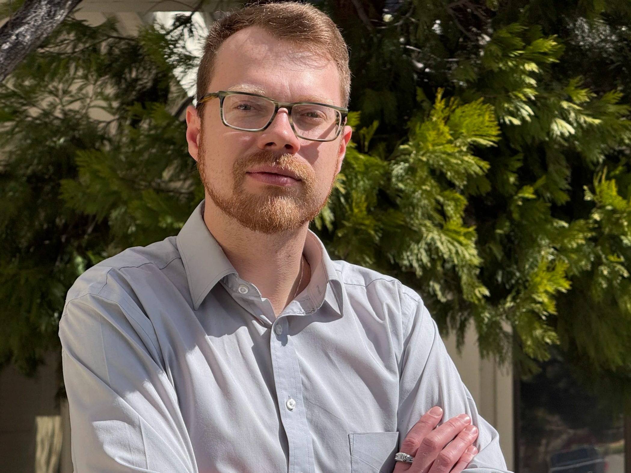 Blaise Jorgensen, candidate for Washoe County Public Administrator, standing outdoors in front of green foliage, wearing a light gray button-up shirt and a ring on his finger.