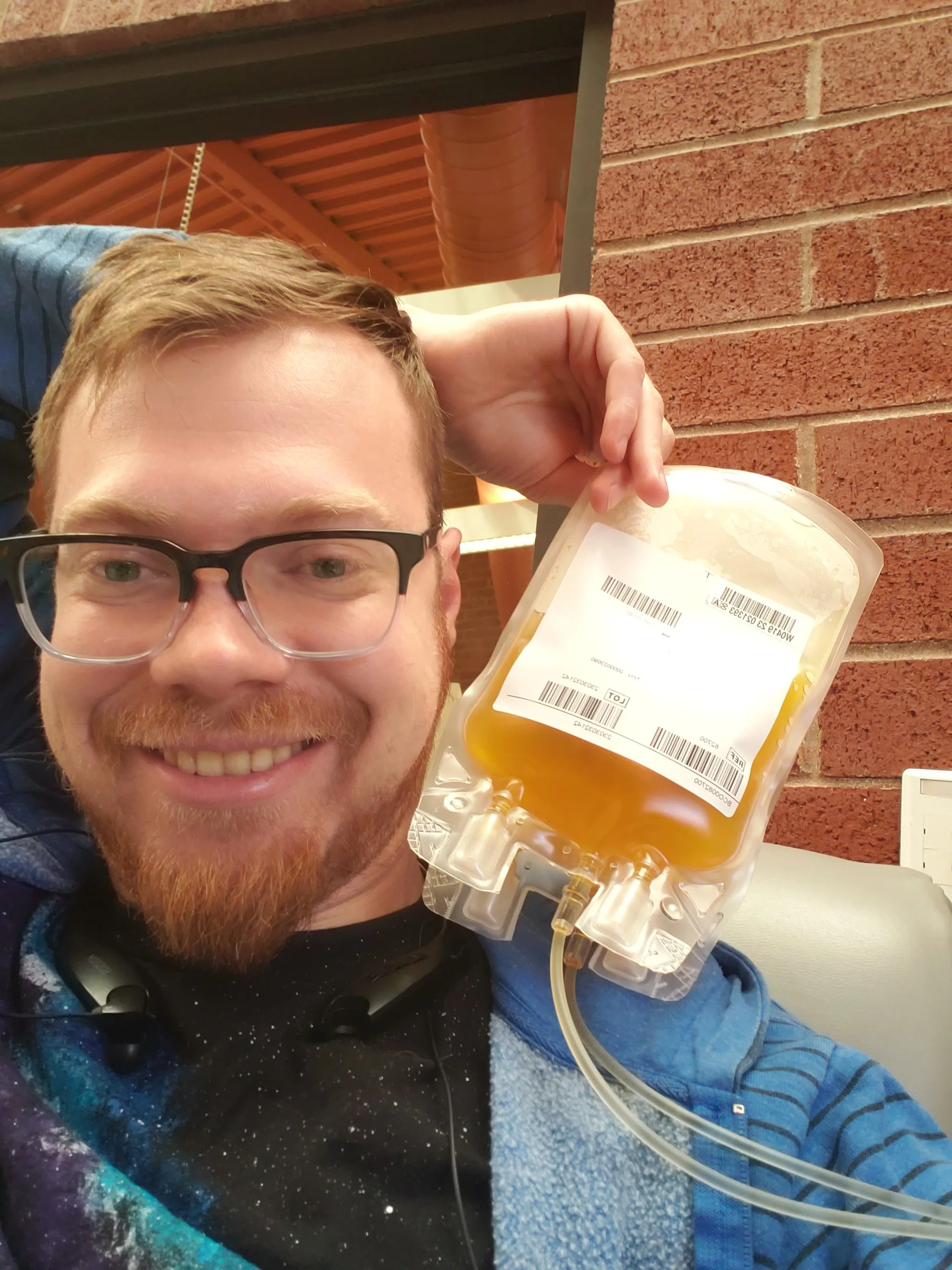Blaise Jorgensen, candidate for Washoe County Public Administrator, holds a blood donation bag filled with blood, sitting in a medical facility with a brick wall in the background.