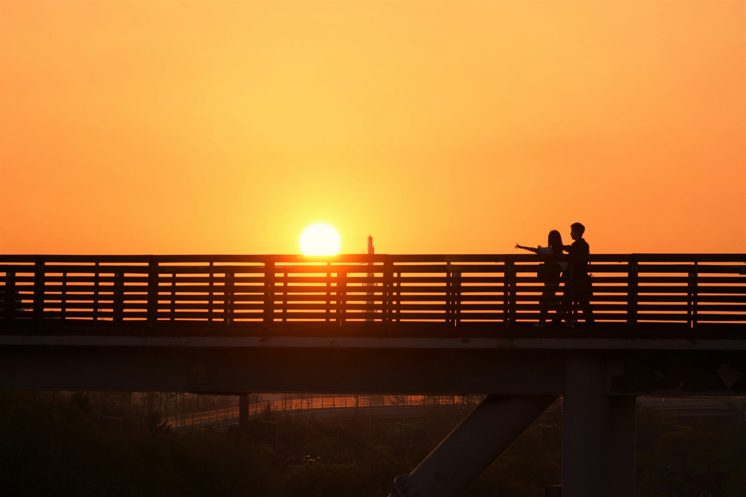 Silhouettes of two people walking together on a bridge during sunset, with a glowing sun in the background and the sky in shades of orange and yellow.