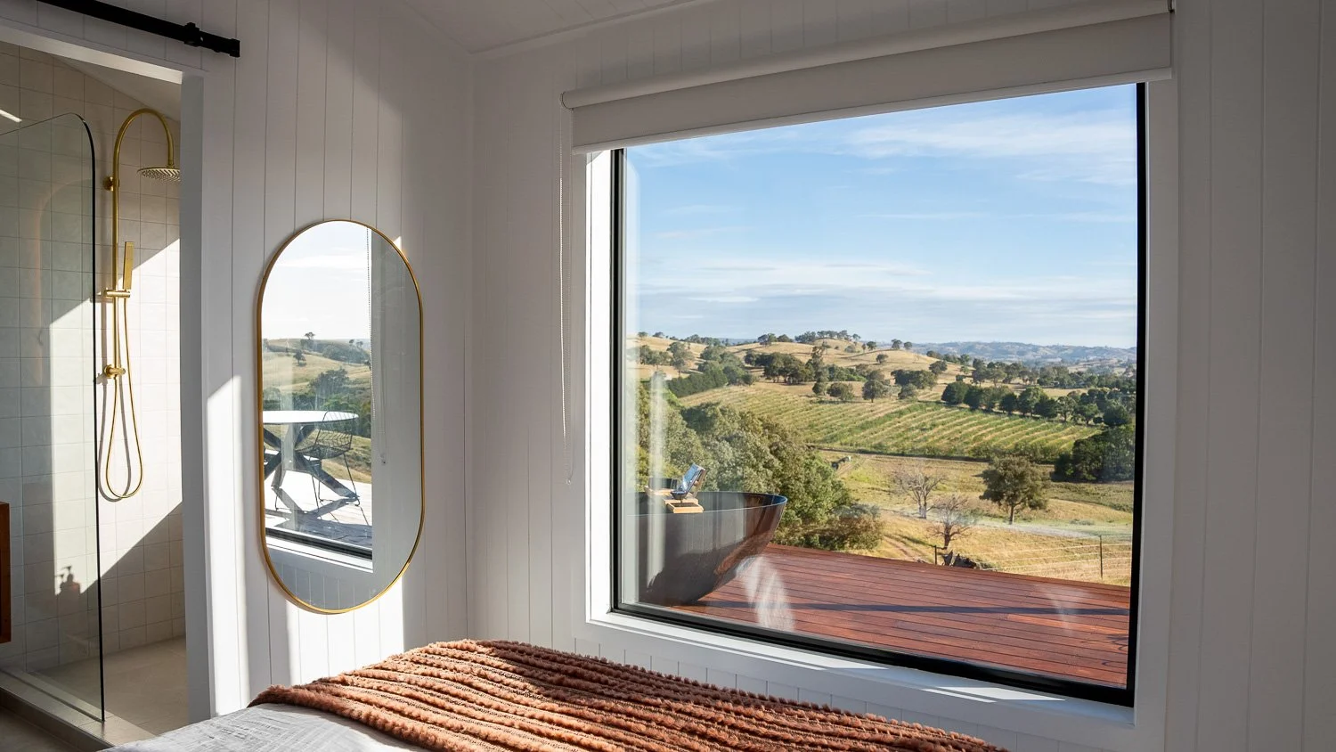 A bedroom with a large window overlooking a scenic view of rolling hills and farmland, including a wooden deck with a hot tub outside.