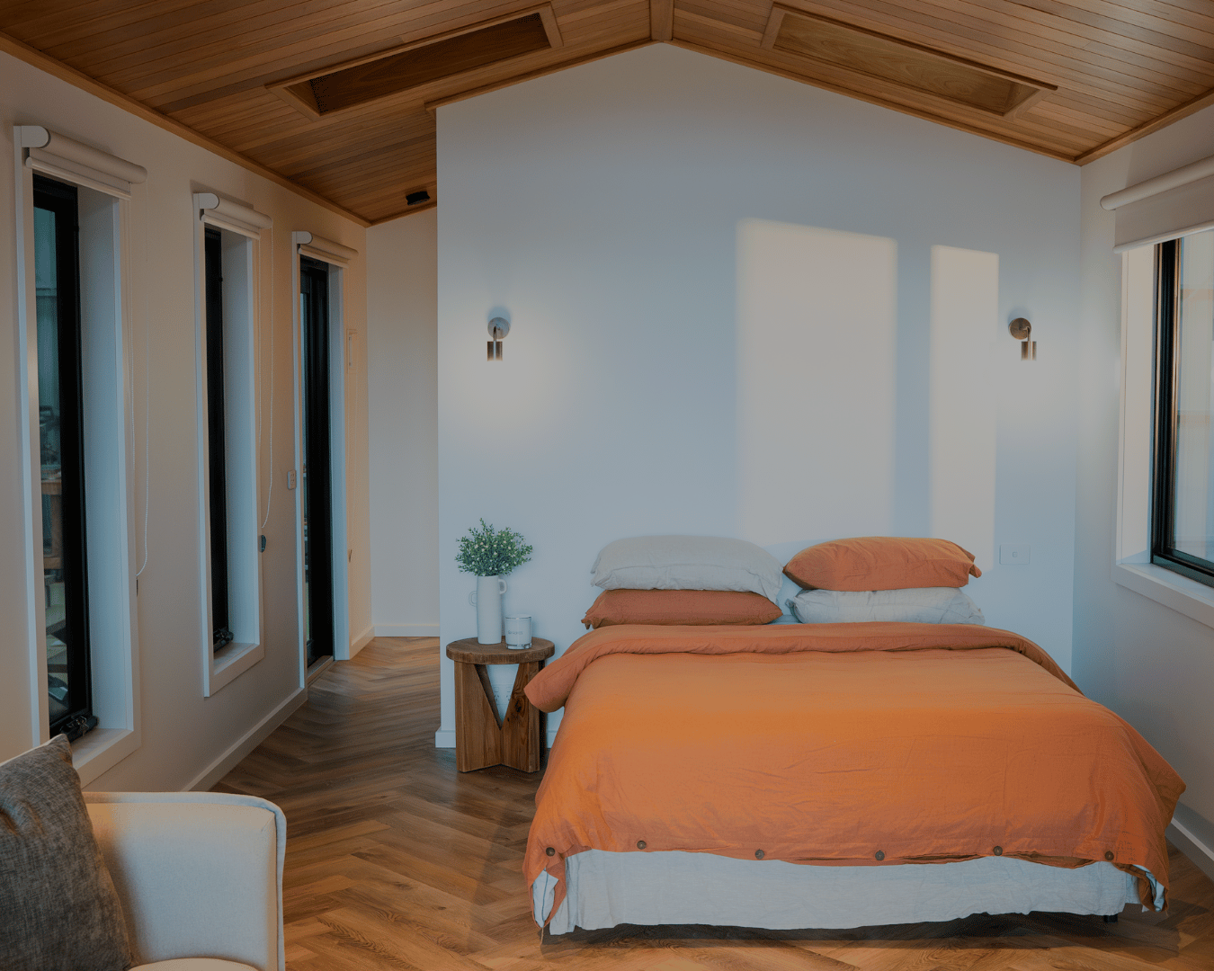 A bedroom with a bed dressed in orange bedding and pillows, a small wooden side table with a vase of greenery and candles, wood-paneled ceiling, light wall sconces, and windows with white blinds.