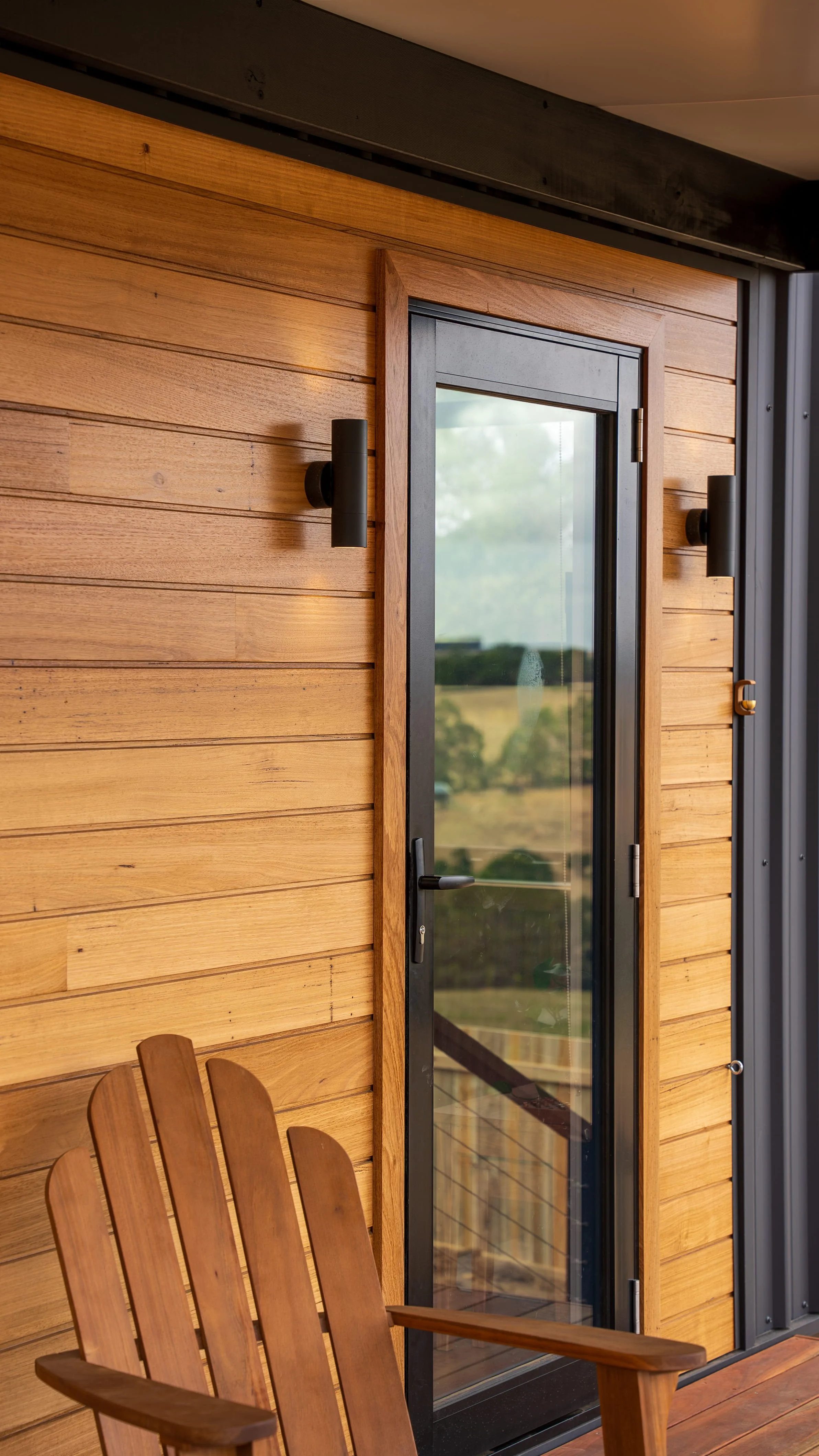 A wooden chair with a curved backrest and armrests in front of a wooden wall, with a glass door revealing a scenic outdoor landscape.