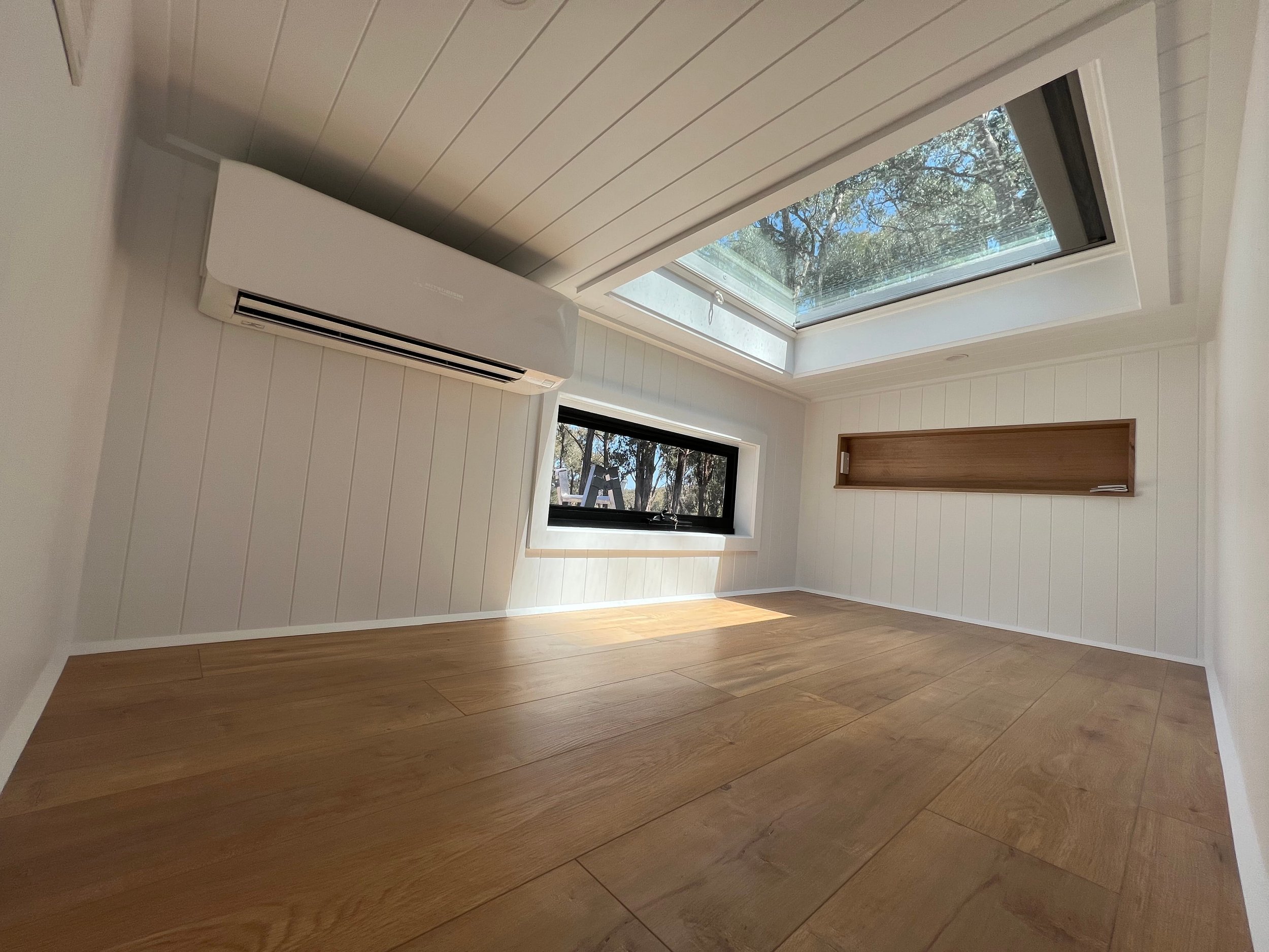A room with white paneled walls, wooden flooring, a skylight window showing trees outside, and a side window, with a wooden shelf on the wall and an air conditioning unit.