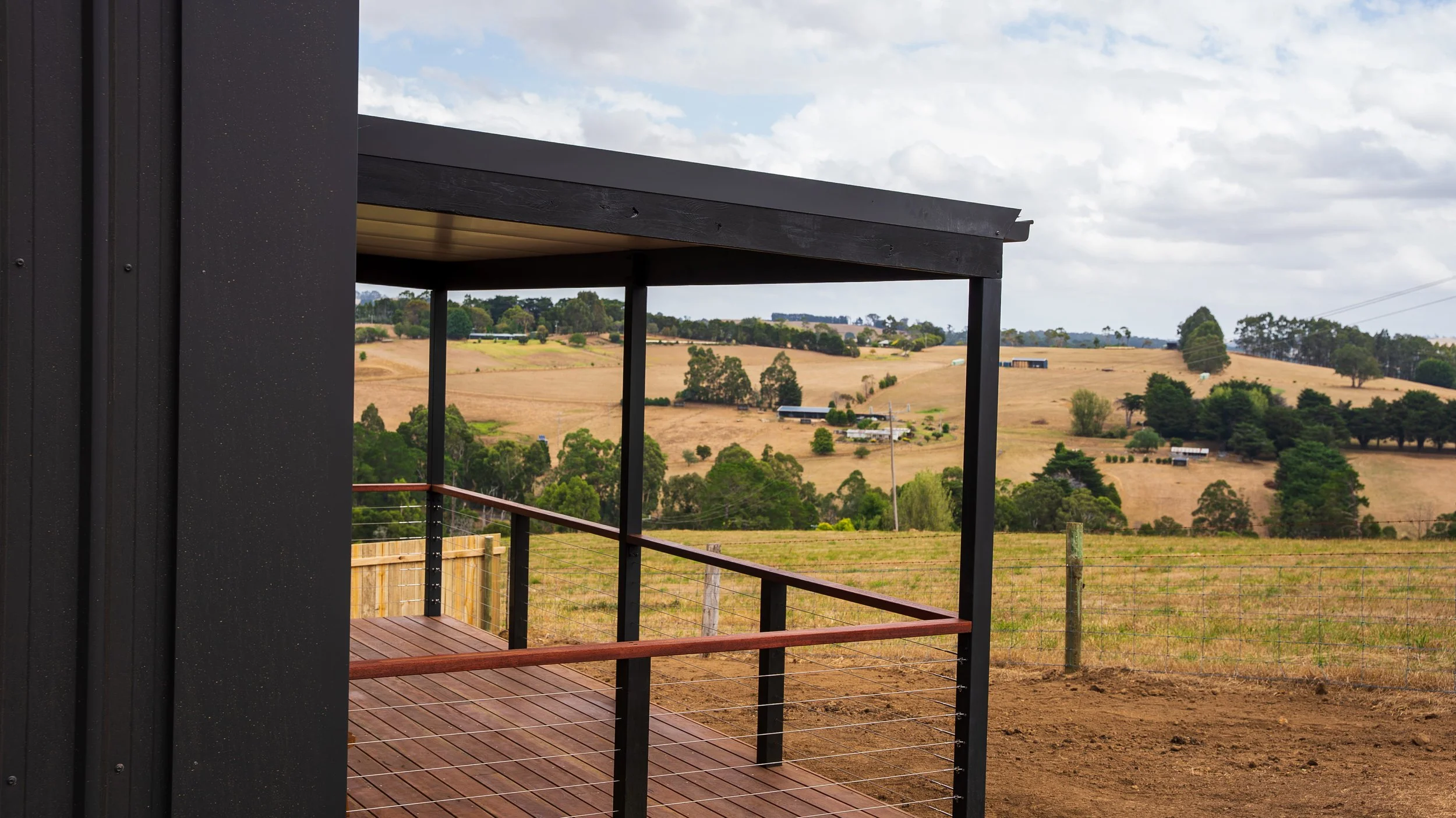 A balcony with black metal framing and wooden flooring overlooks a rural landscape with rolling hills, scattered trees, and farm buildings under a partly cloudy sky.