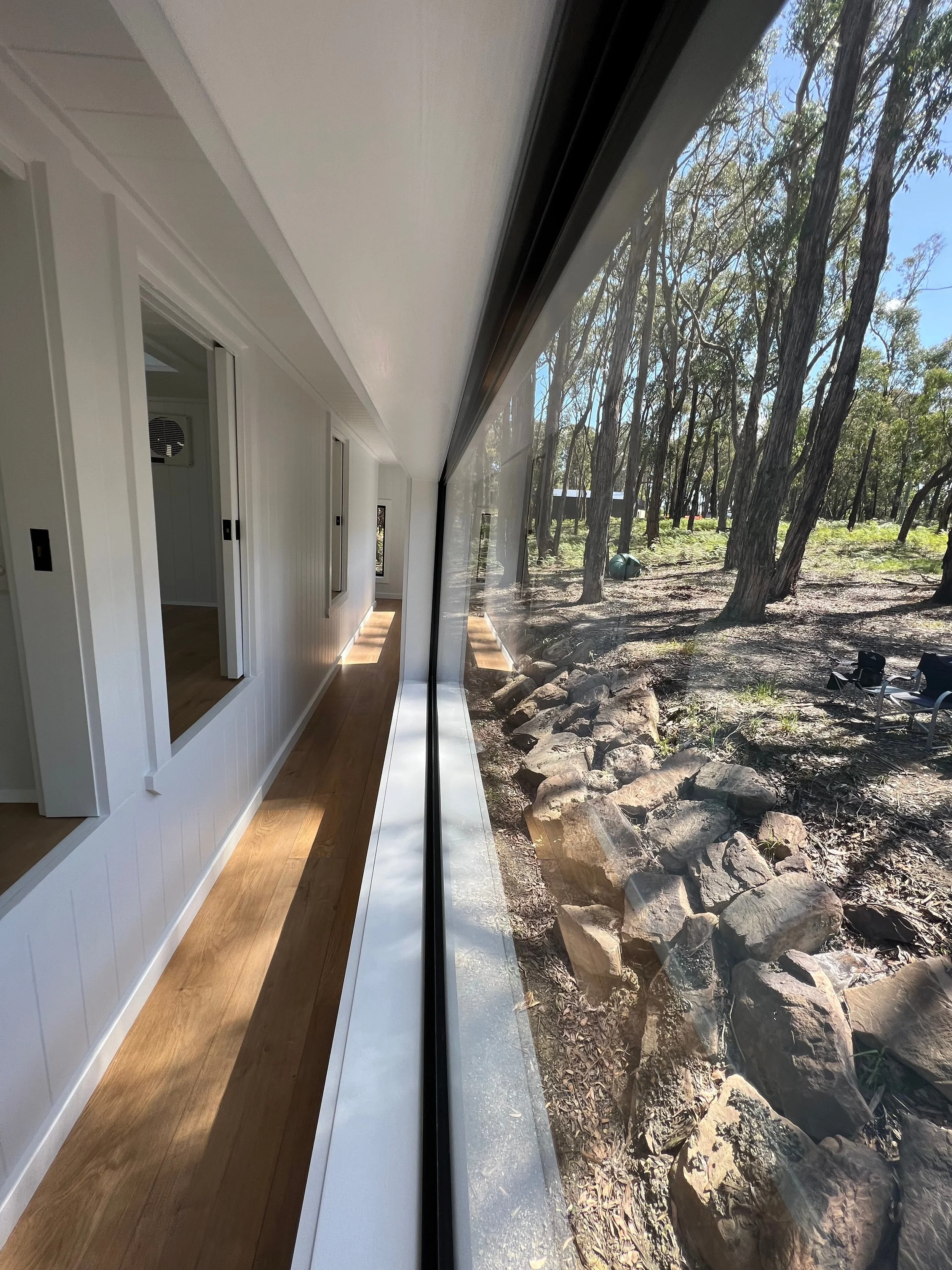 Interior of a modern house with white walls, wooden floors, and large glass windows that overlook a wooded outdoor area with trees and rocks.