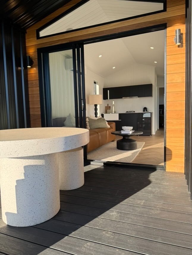 View from an outdoor deck showing a round concrete table and an open sliding door leading into a modern living room with a tan sofa, black coffee table, and a kitchen with black cabinets.