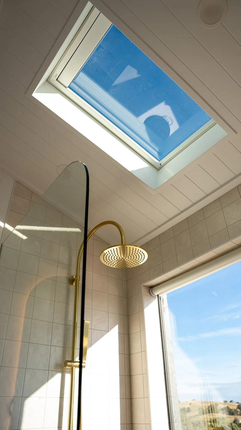 Bathroom with a skylight, a shower with a gold showerhead, a glass shower partition, and a large window showing a landscape outside.