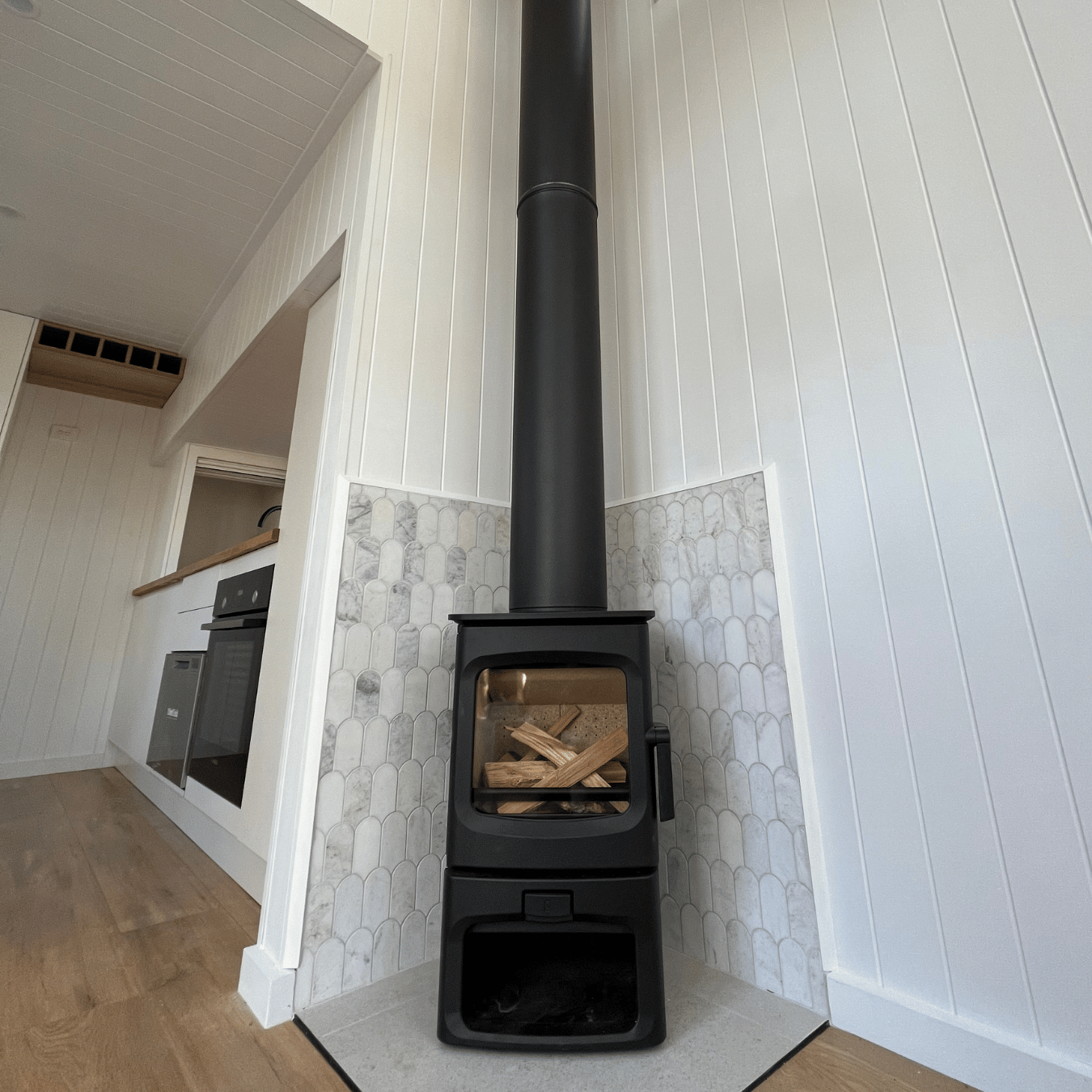 A black wood stove with a glass door filled with firewood, situated in a corner with a tall black chimney pipe extending upward. The stove is next to white paneled walls with marble-like tiling around it, inside a cozy, modern home.