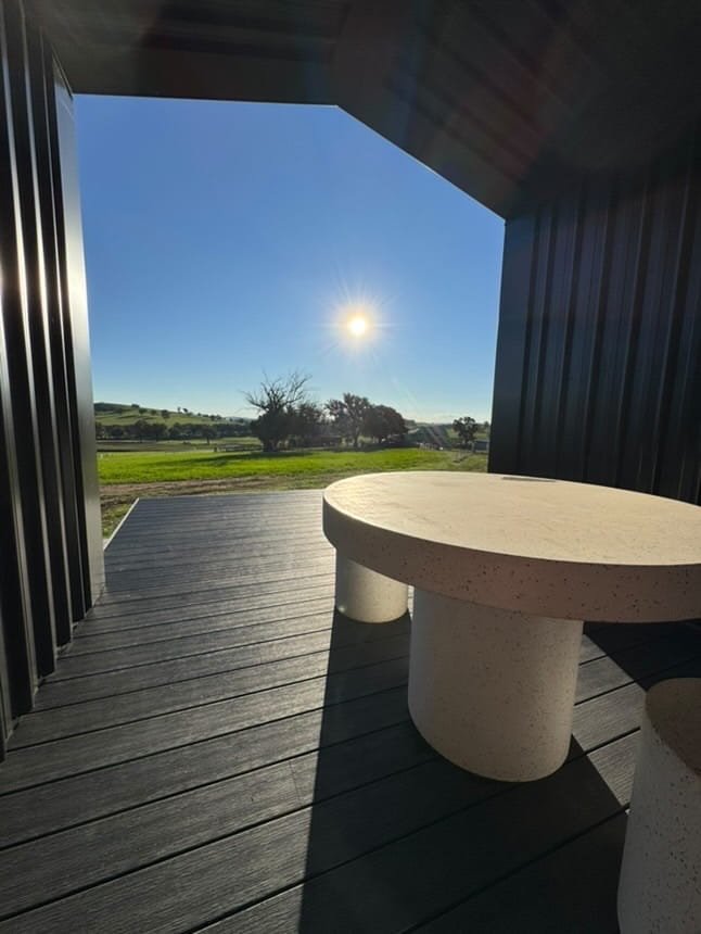 View from a modern covered porch with black siding, overlooking a green field and trees under a clear blue sky with the sun shining.