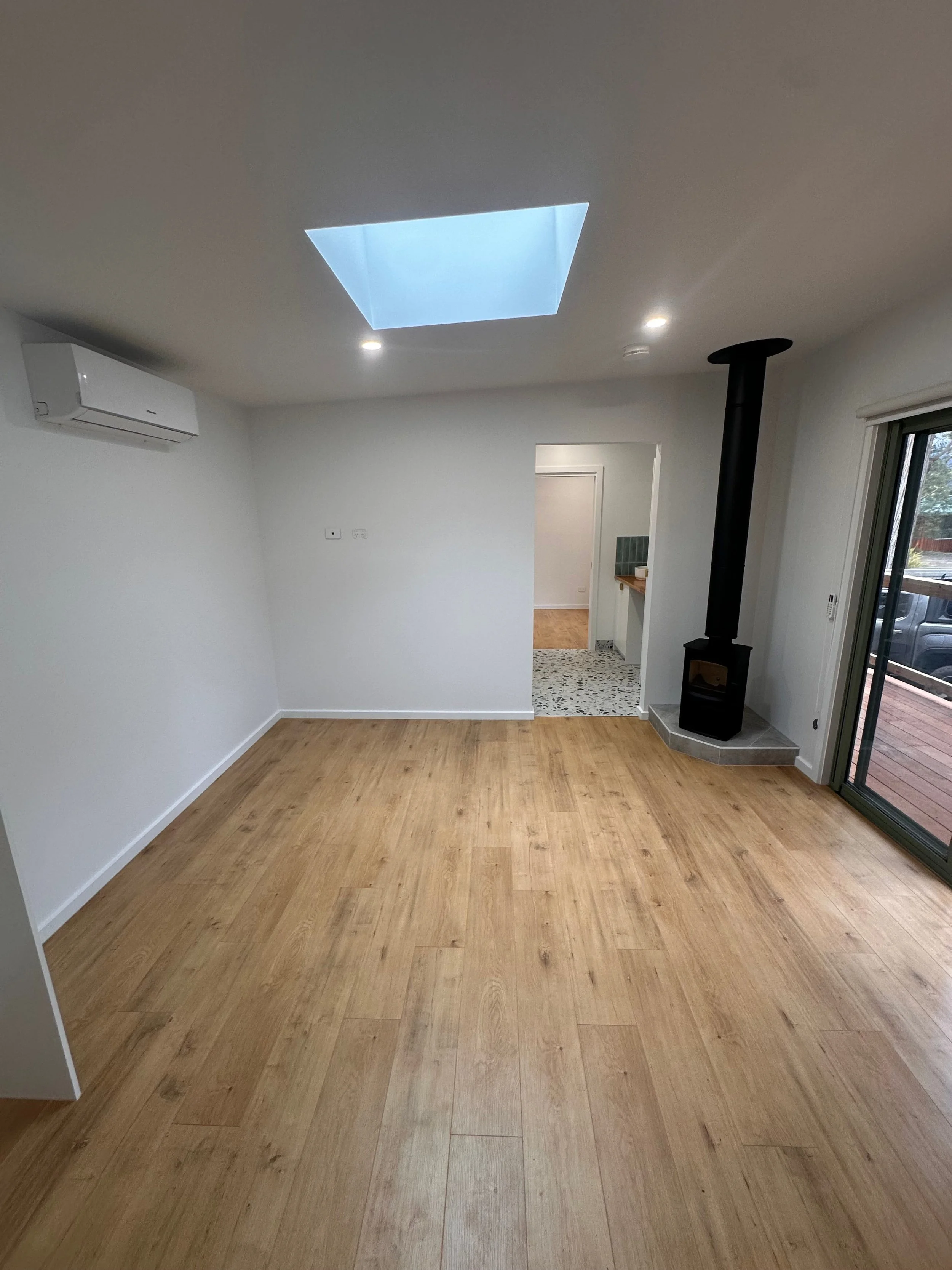 Empty living room with wooden floor, white walls, a wood stove, air conditioning unit, and sliding glass door to an outdoor deck.