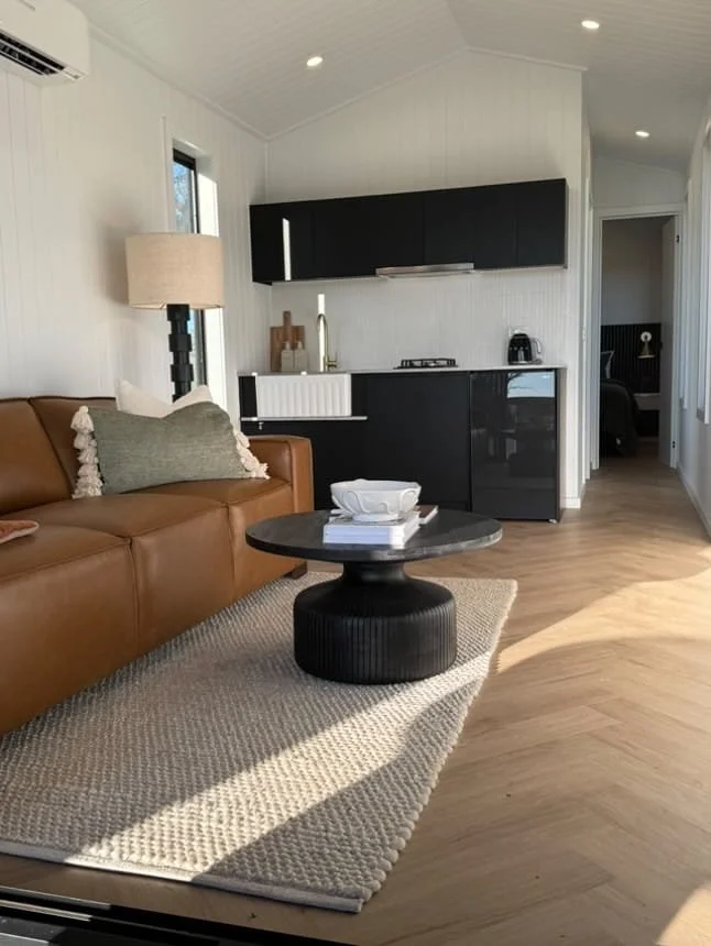 Living room with a brown leather sofa, a black round coffee table, a beige area rug, a white wall with a window, a lamp, and a kitchen area with black cabinets and a small black refrigerator.