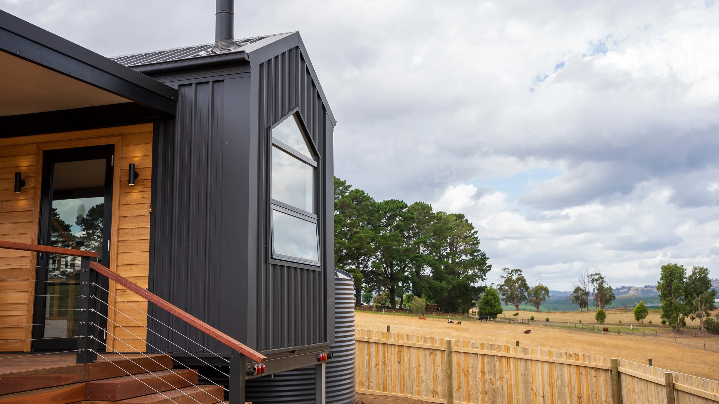 A modern tiny home with black metal siding, wooden accents, and large windows, set in a rural area with a wooden fence, trees, and grazing animals under a cloudy sky.