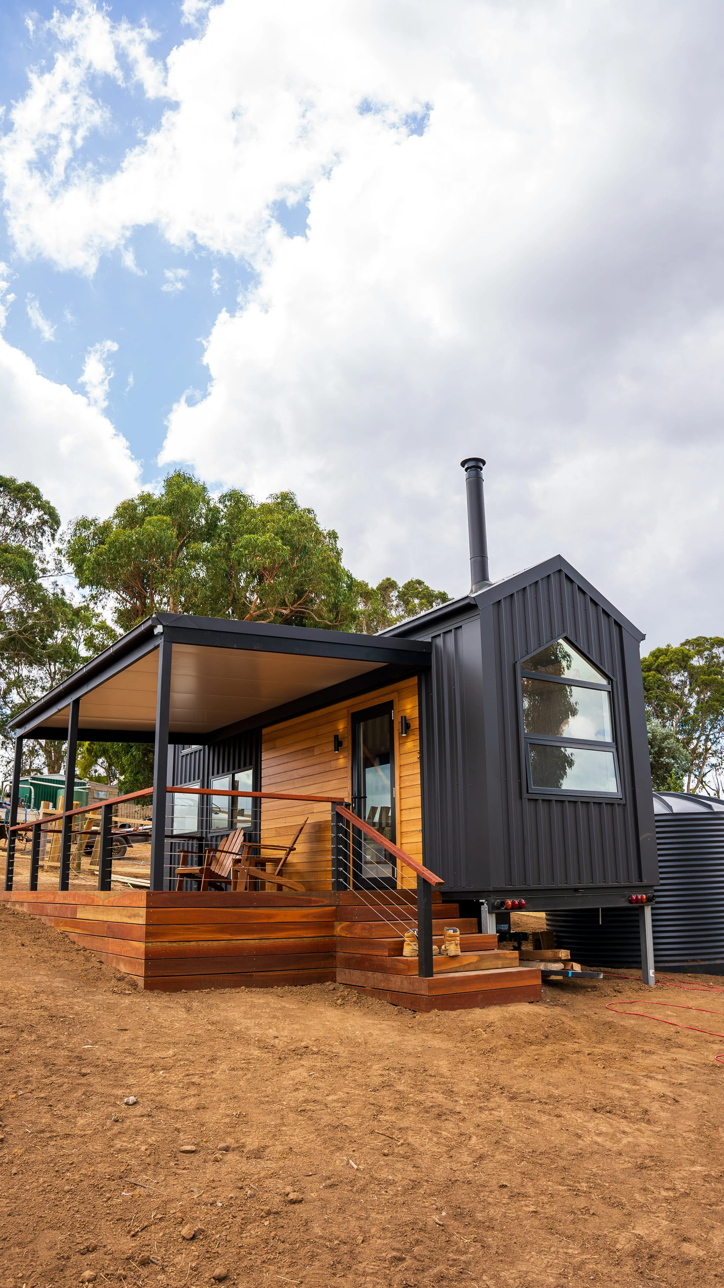 Arnhem design tiny house with black metal exterior and large windows, surrounded by trees and open sky, featuring a wooden deck with outdoor seating.