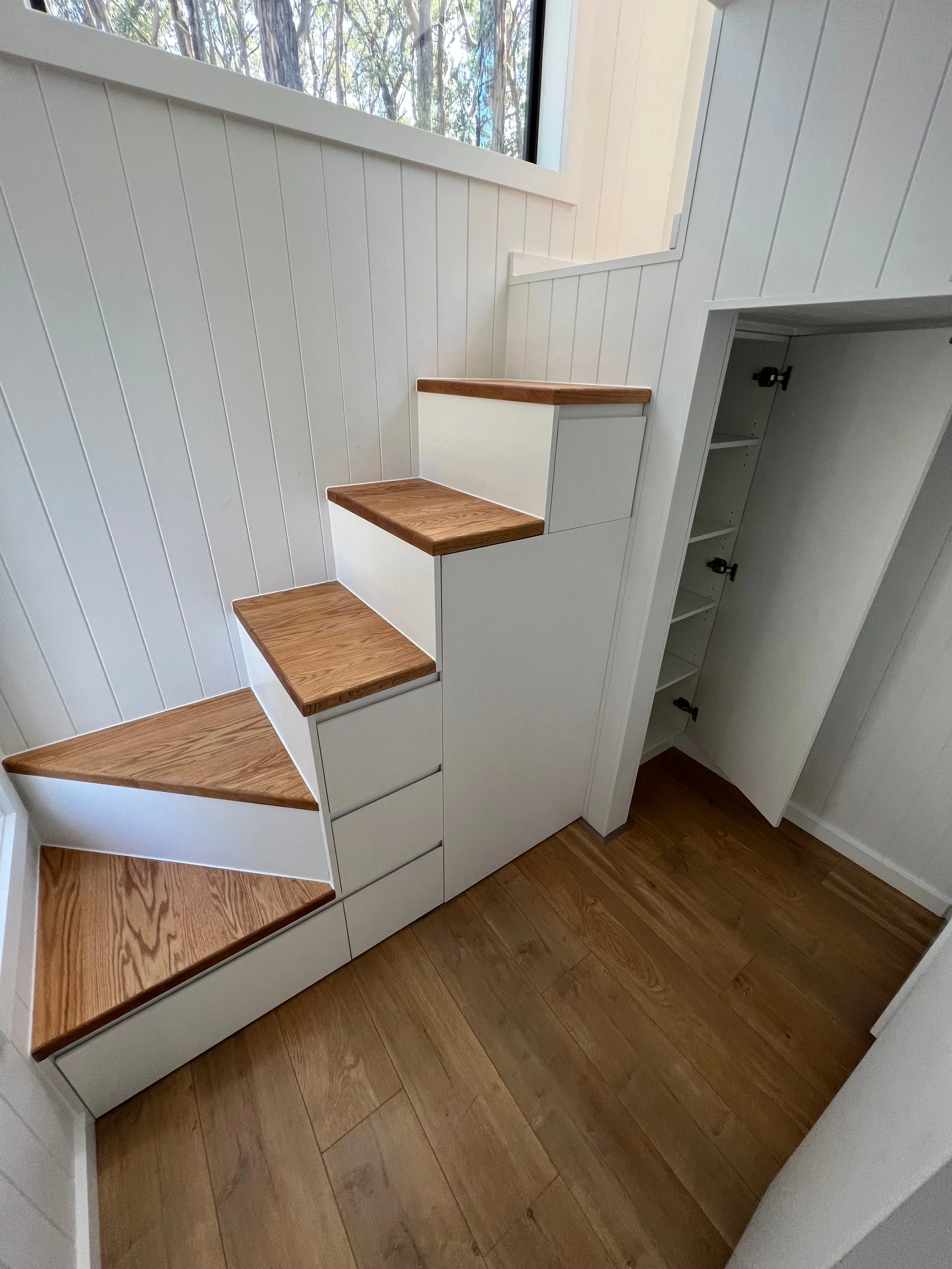Interior view of a staircase with wooden steps, white risers, and white wall paneling, next to a large window showing trees outside.