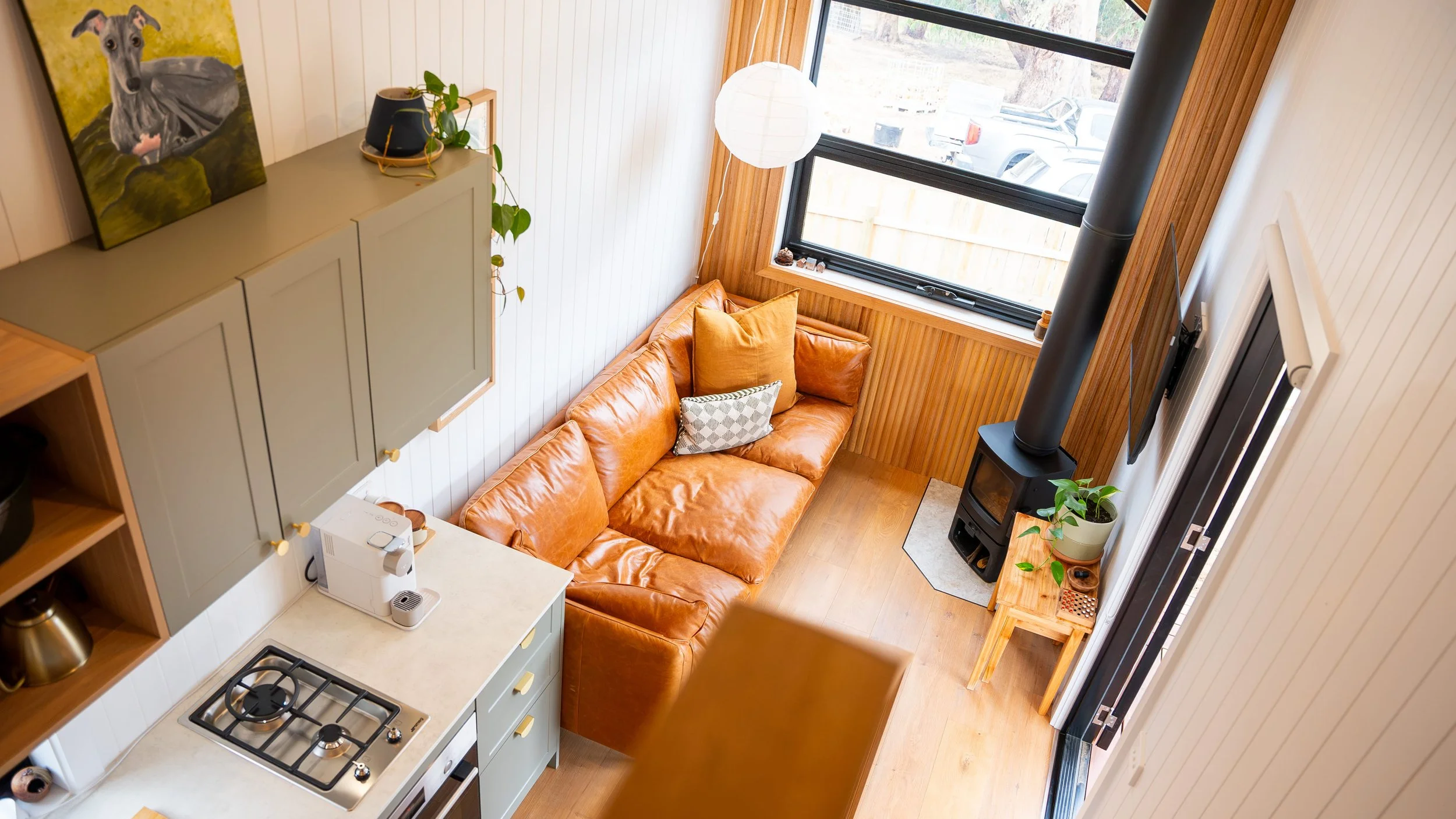 Interior view of a cozy living room with a leather couch, large window, wood stove, potted plants, and wall art, seen from above.