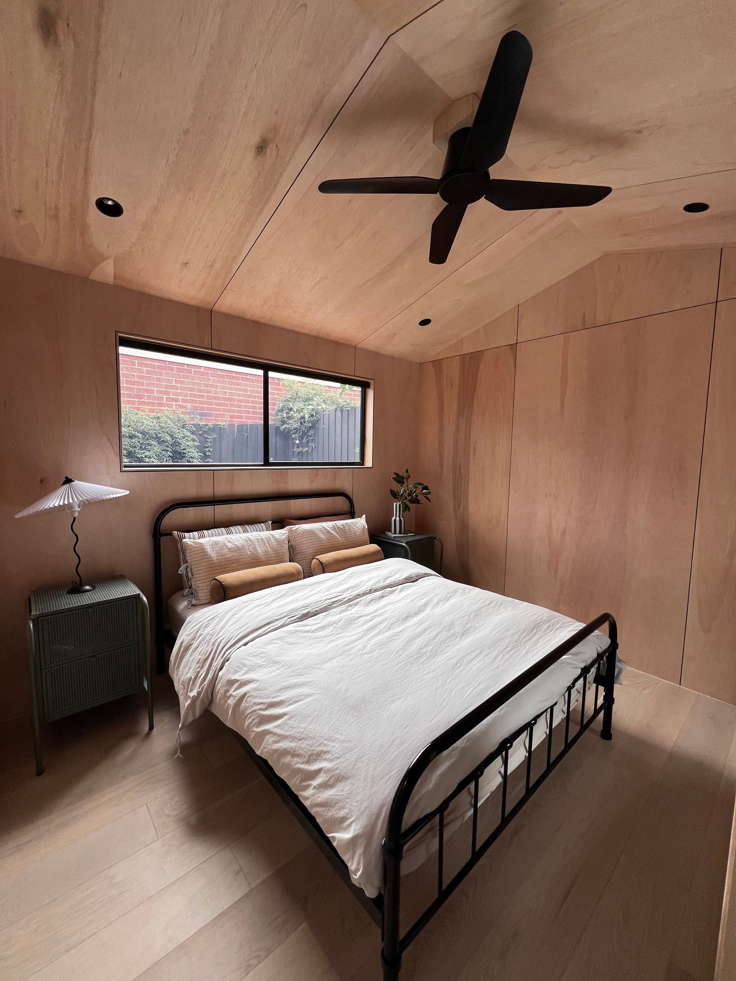 A bedroom with light wooden walls and ceiling, a bed with white bedding, and a window showing a brick wall and some greenery outside.