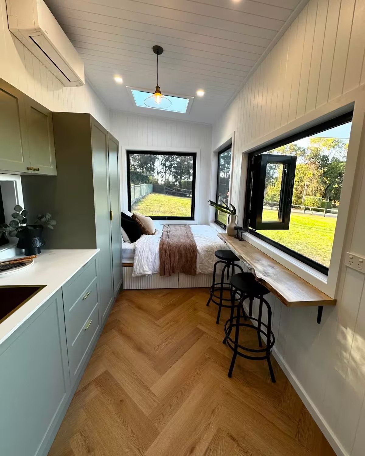 Small bedroom with a bed under large windows, kitchen kitchenette to the left, and a wooden bar with two black stools to the right, overlooking a green yard.