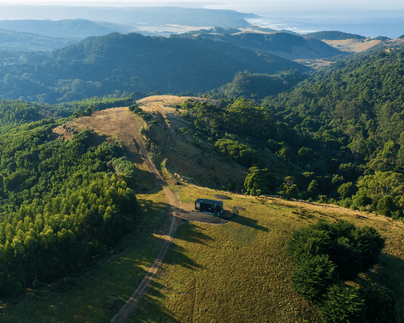 Aerial view of green, hilly landscape with a black bus parked on a dirt path, overlooking forested hills and distant coastline.