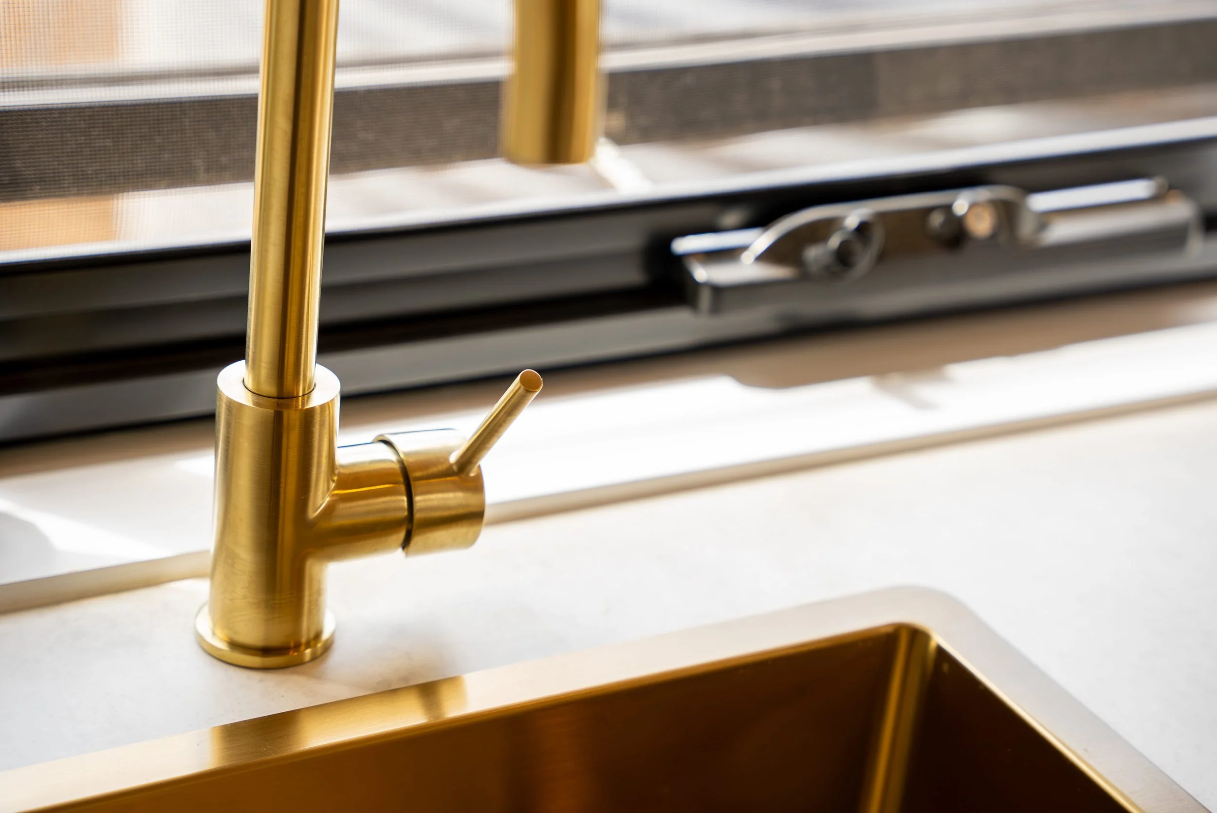 Close-up of a modern kitchen sink with a gold-colored faucet and a gold pop-up drain, set in a white countertop, with a window screen behind the sink.