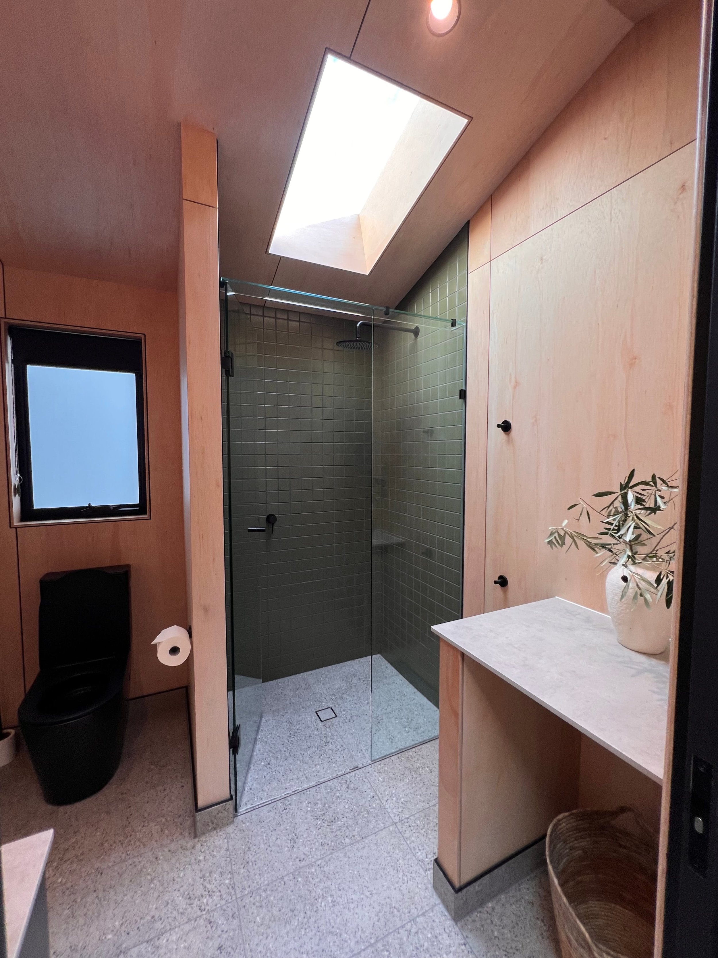 Modern bathroom with tiled walk-in shower, skylight, black toilet, and wooden cabinetry with a white countertop and plant