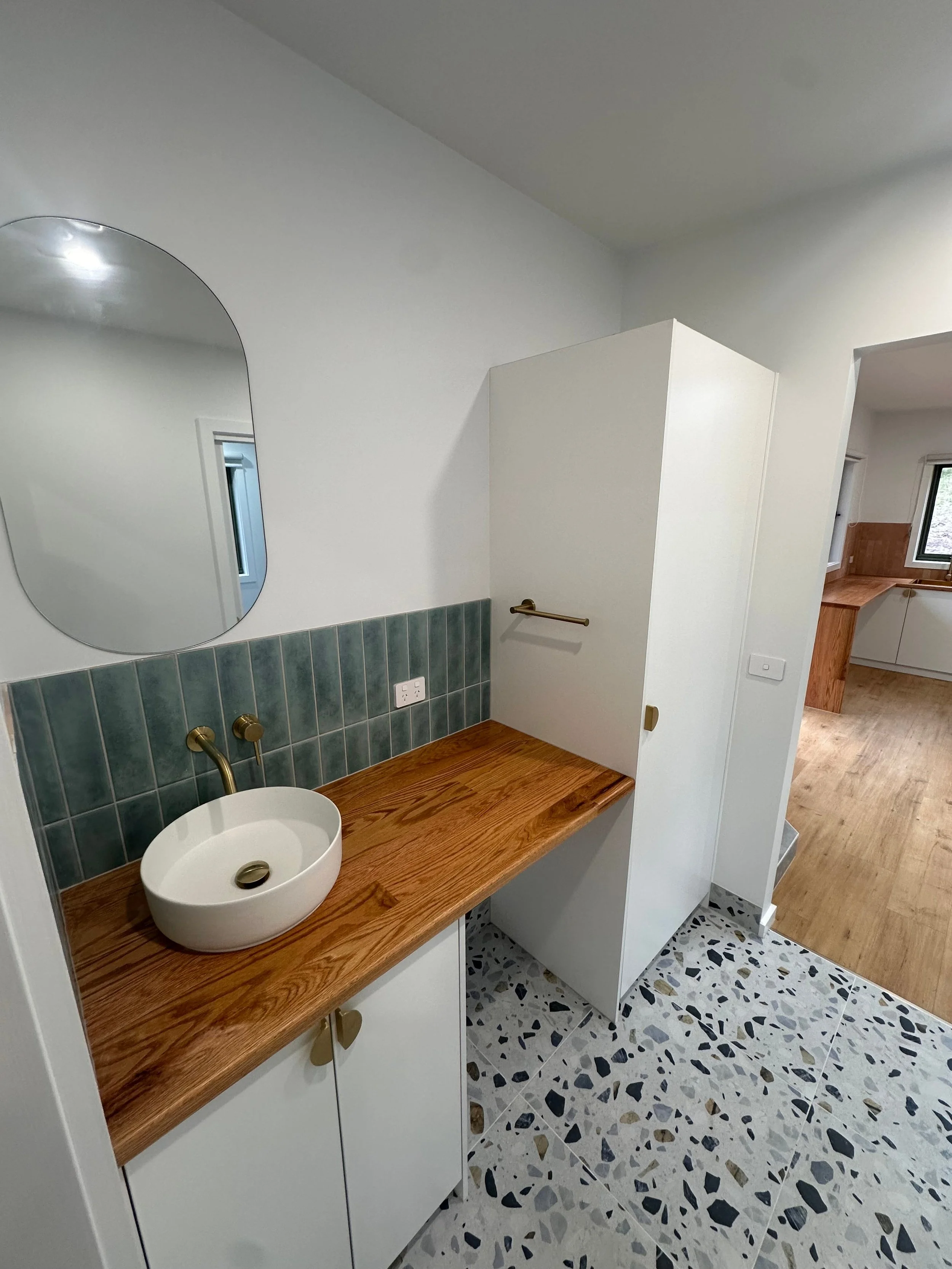 A bathroom with a wooden vanity featuring a white vessel sink and brass fixtures, a framed mirror, green tiled wall, white cabinets, a partial shower stall door, and a private kitchen area in the background with wooden cabinetry and window.