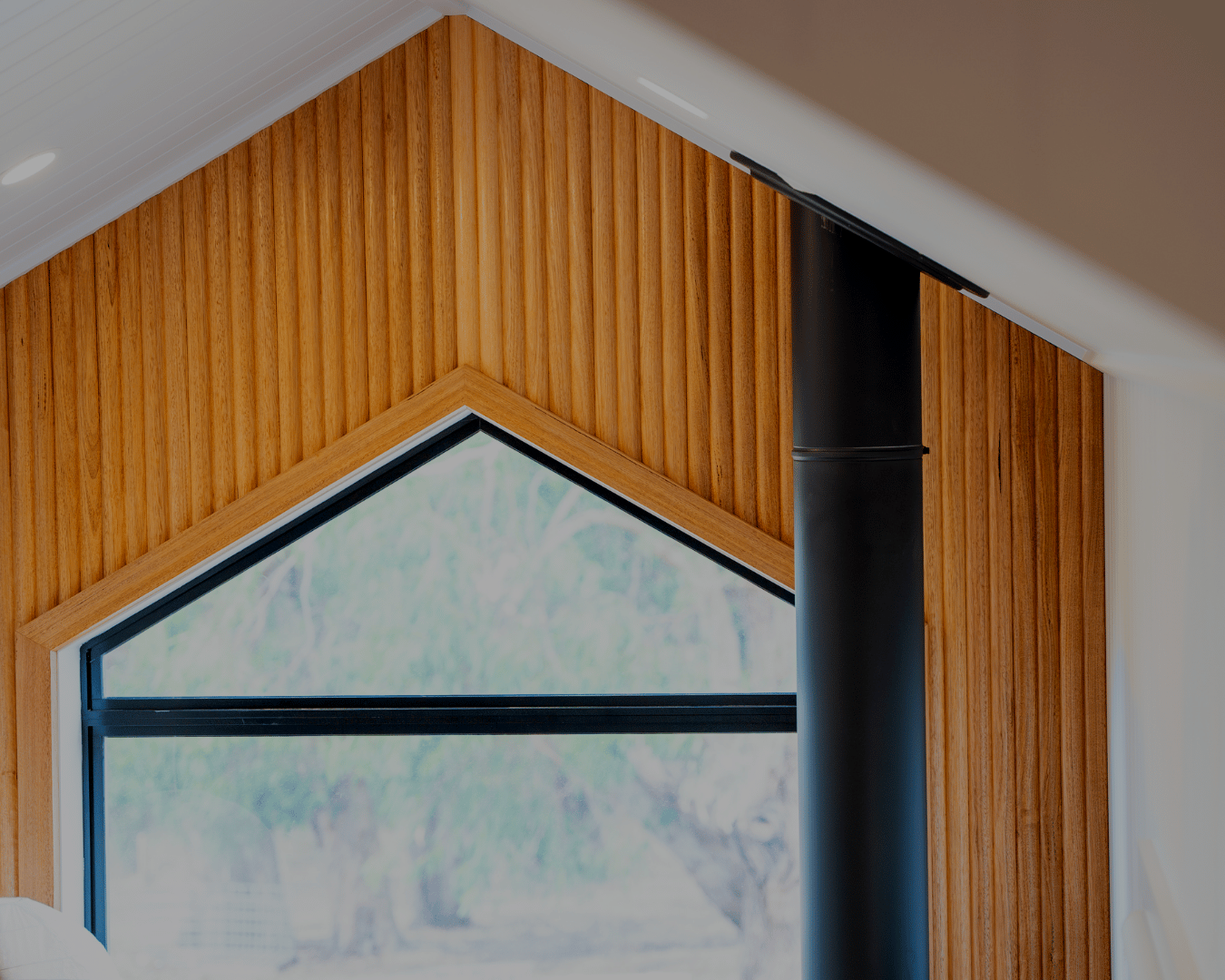 Close-up of an interior corner of a modern house with wood paneling, a large triangular window, and a black chimney or pipe.