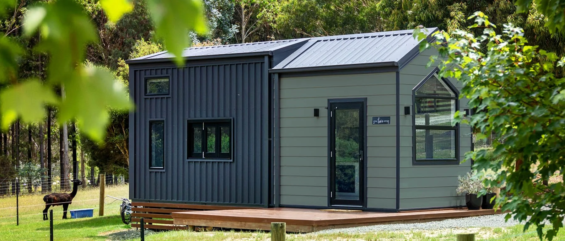 A modern tiny house with black and gray exterior, located in a green, wooded area, with a wooden deck and potted plants outside.