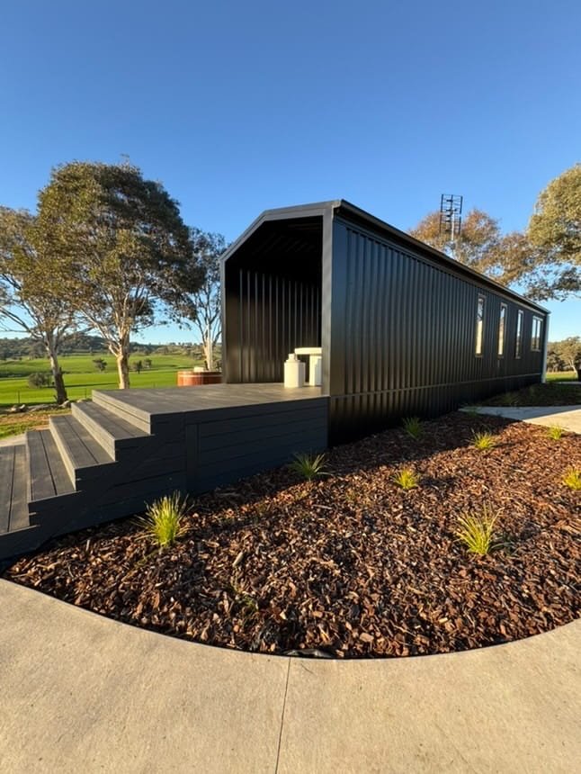 A tiny black house in a park with trees in the background, wooden stairs leading up to it, and a concrete pathway in front.