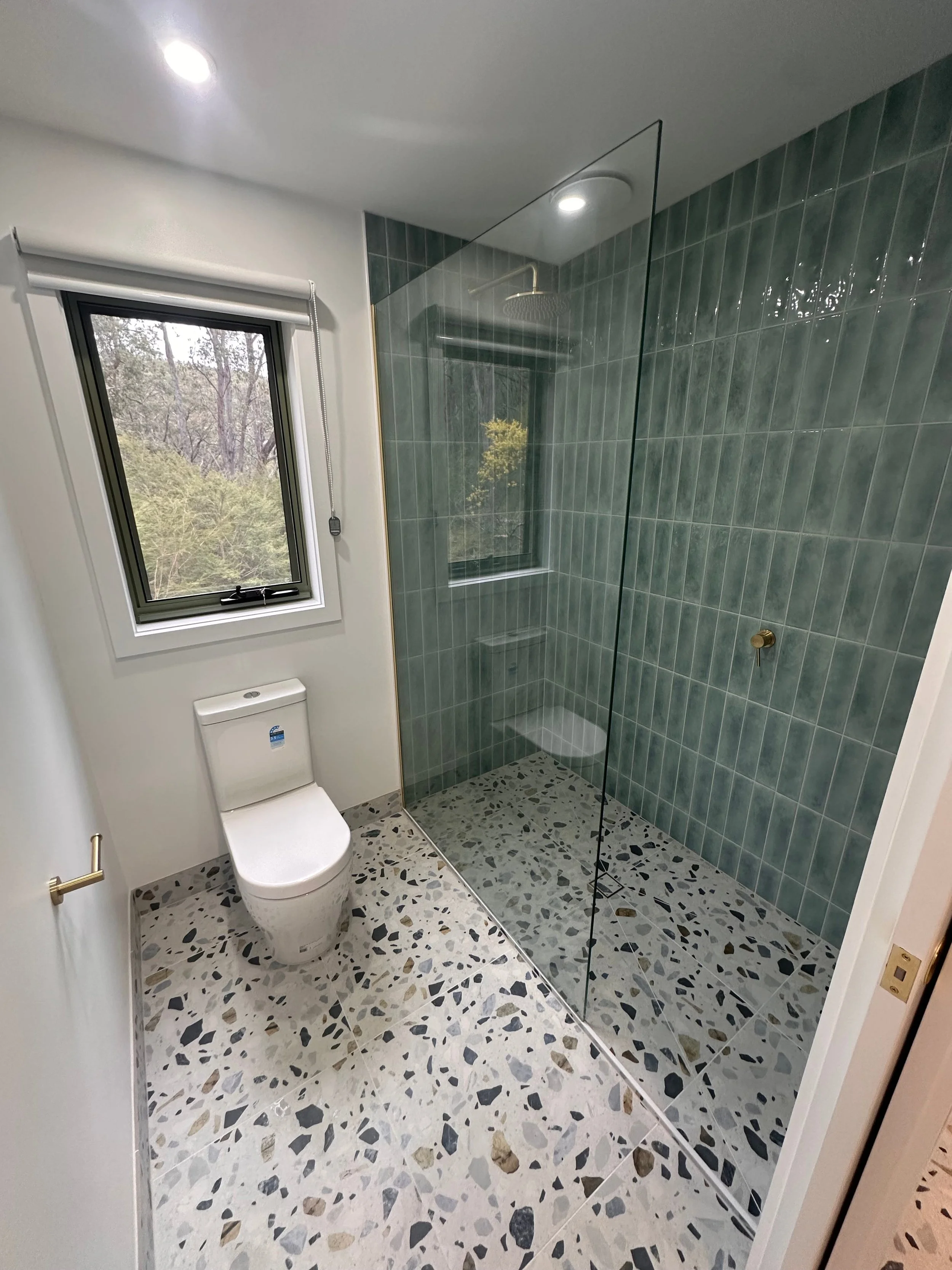Modern bathroom with terrazzo floor, a white toilet, black framed window with a view of trees, and a glass shower enclosure with green tiles.