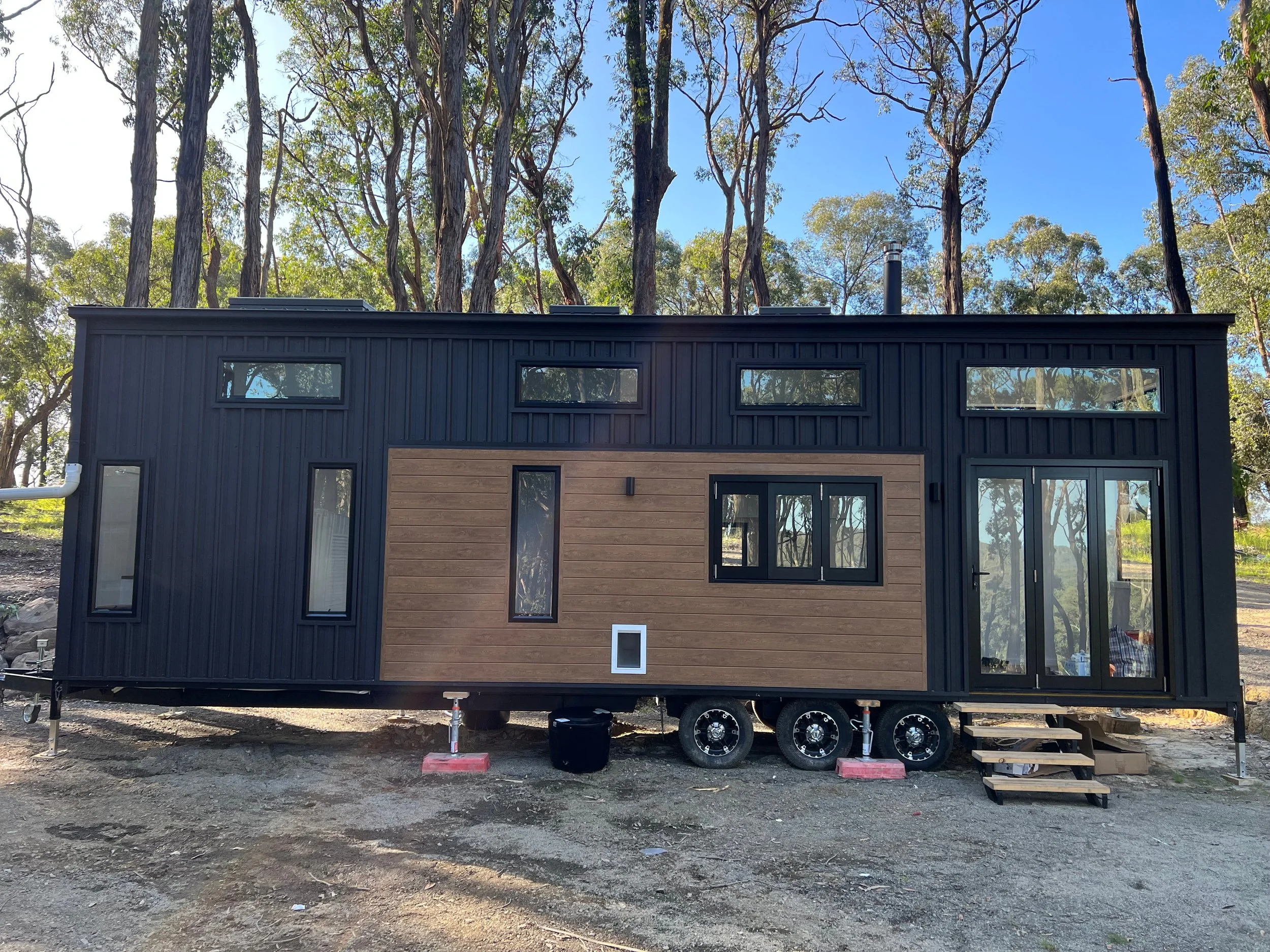 A modern tiny house on wheels with black metal siding and wood paneling. It has several narrow rectangular windows and a sliding glass door. The house is elevated on a trailer with wheels and is situated in a wooded area with tall trees in the backgr
