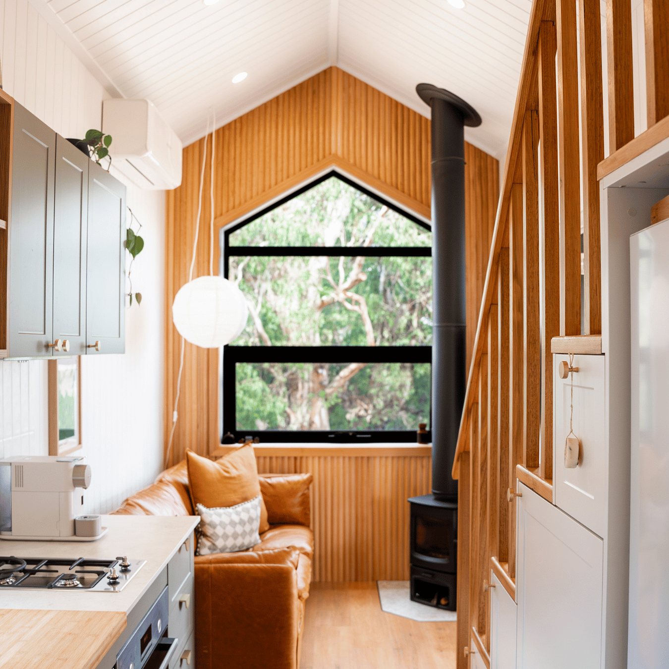 Interior of a cozy, modern living space with a sloped ceiling, large triangular window showing trees outside, a black metal chimney, a leather sofa with cushions, kitchen cabinets, and a white lamp, with wooden accents and natural light.