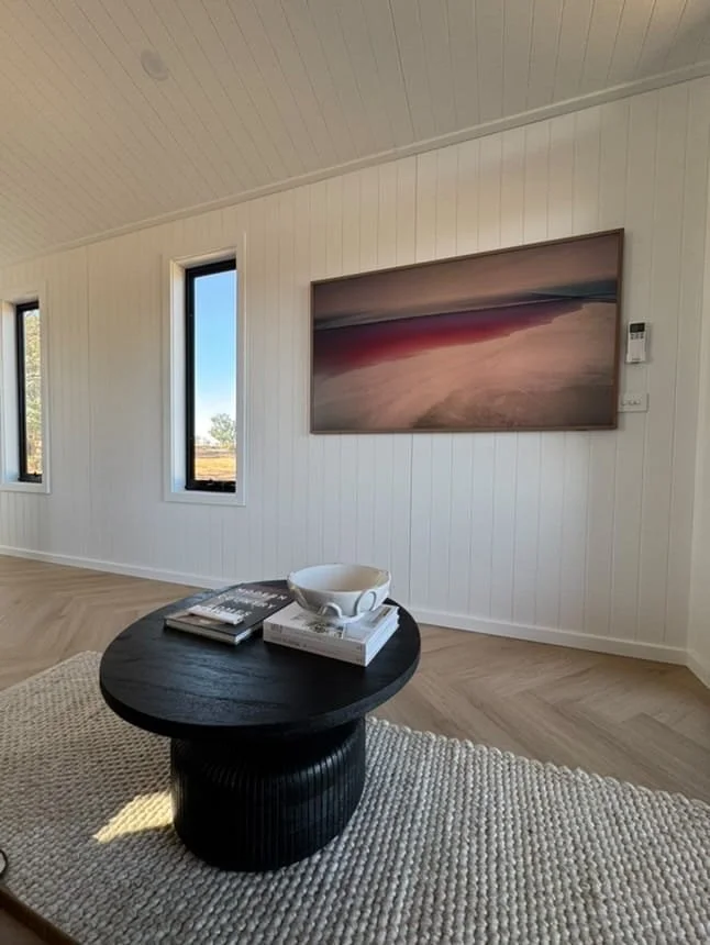Interior of a room with white paneled walls, three narrow windows showing outside trees and sky, and a large abstract landscape painting on the wall. In the foreground, a black round coffee table with a textured cylindrical base holds a white bowl an