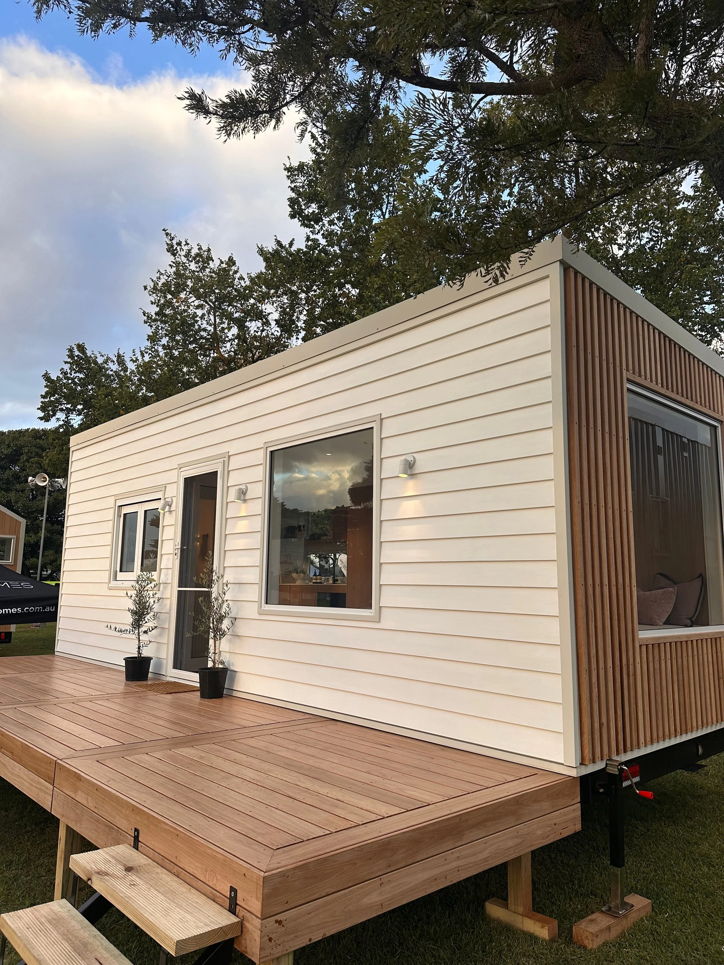 A tiny house on a trailer with a wooden deck in front, white siding, large windows, and a small outdoor seating area, surrounded by green trees under a partly cloudy sky.