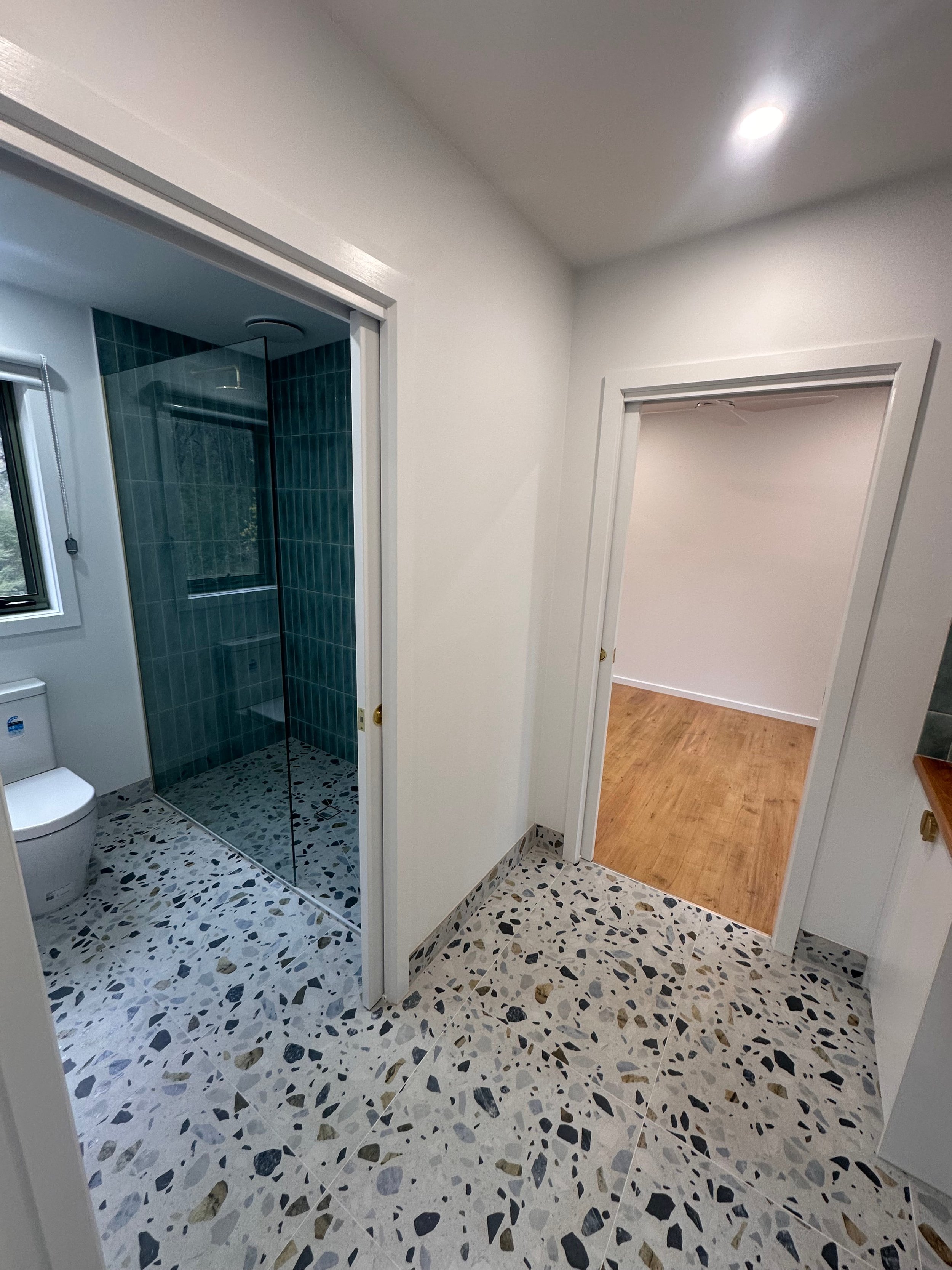 A bathroom with a shower area that has dark green tiles and a glass door, a white toilet, and a window. There is an adjoining room with light-colored wooden flooring visible through an open doorway. The bathroom floor has a terrazzo pattern with blac