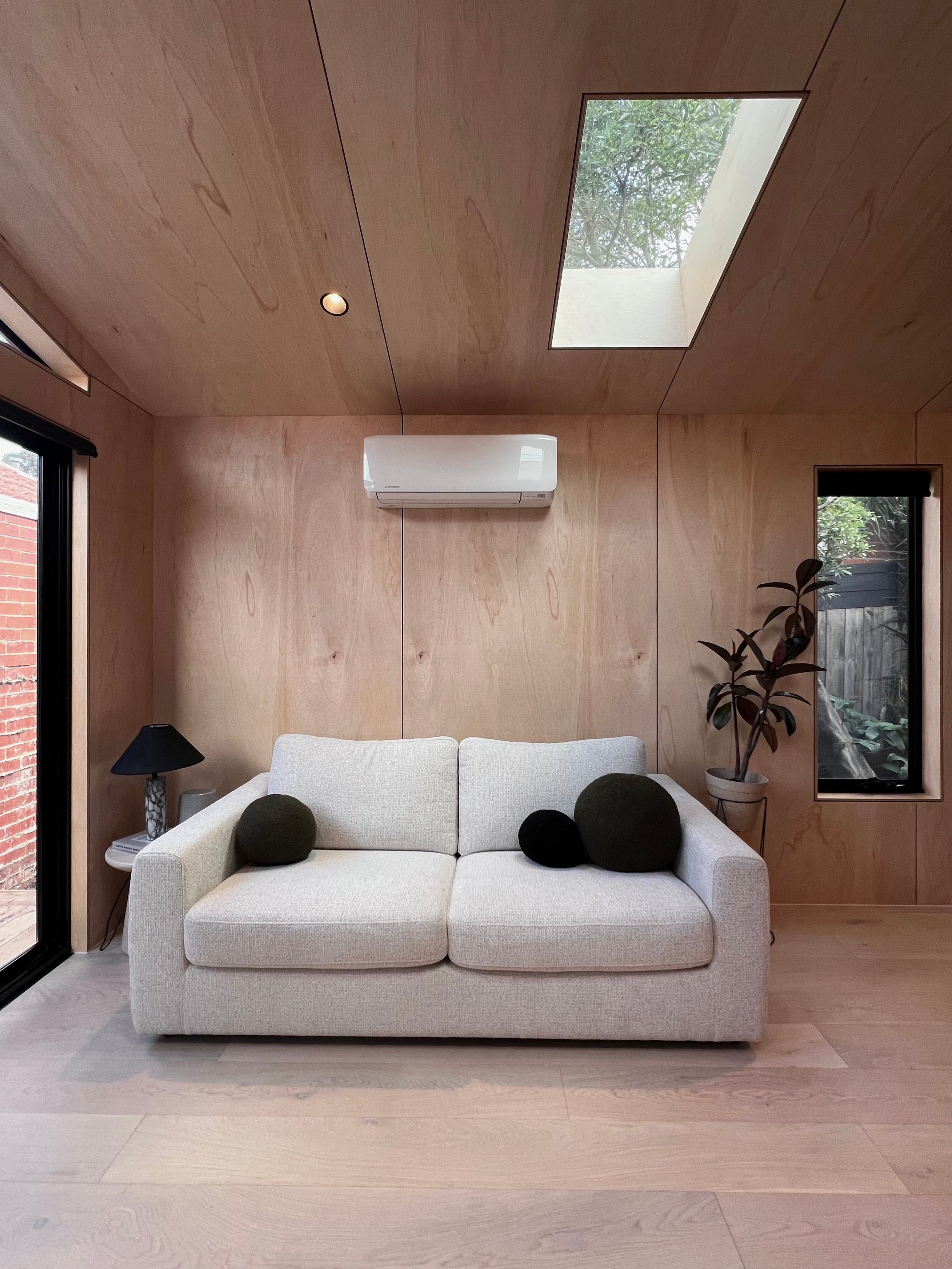 A modern living room with a white sofa, dark green pillows, a side table with a black lamp, a potted plant, windows, and a wooden wall with a skylight.