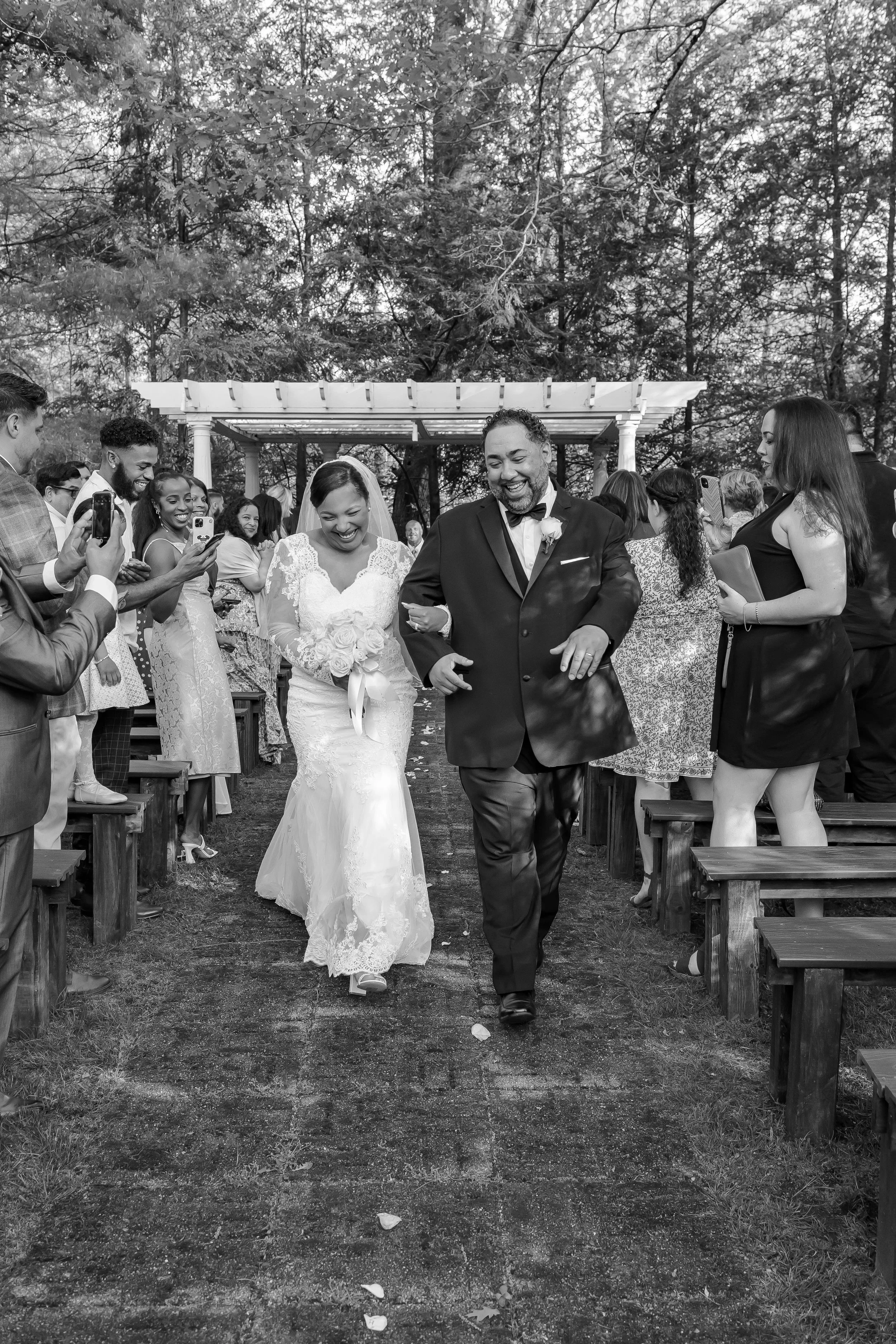 Black and white photo of a bride and groom walking down the aisle at an outdoor wedding ceremony, smiling and surrounded by guests taking pictures.