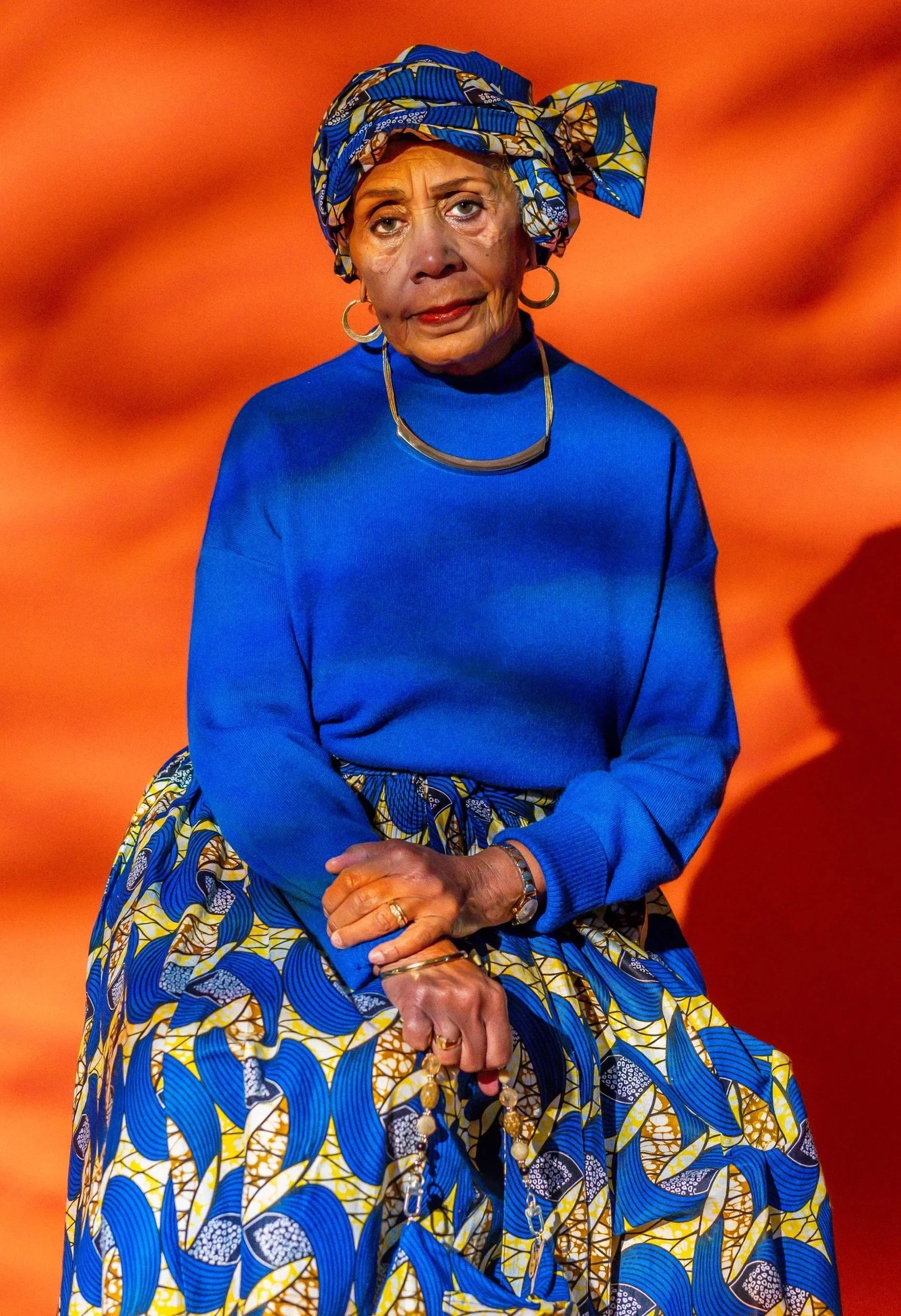 An elderly woman wearing a vibrant blue sweater, a matching colorful patterned skirt, and a headwrap with blue, yellow, and black patterns. She has gold hoop earrings, a gold necklace, and is sitting with her hands clasped. The background is a blurre