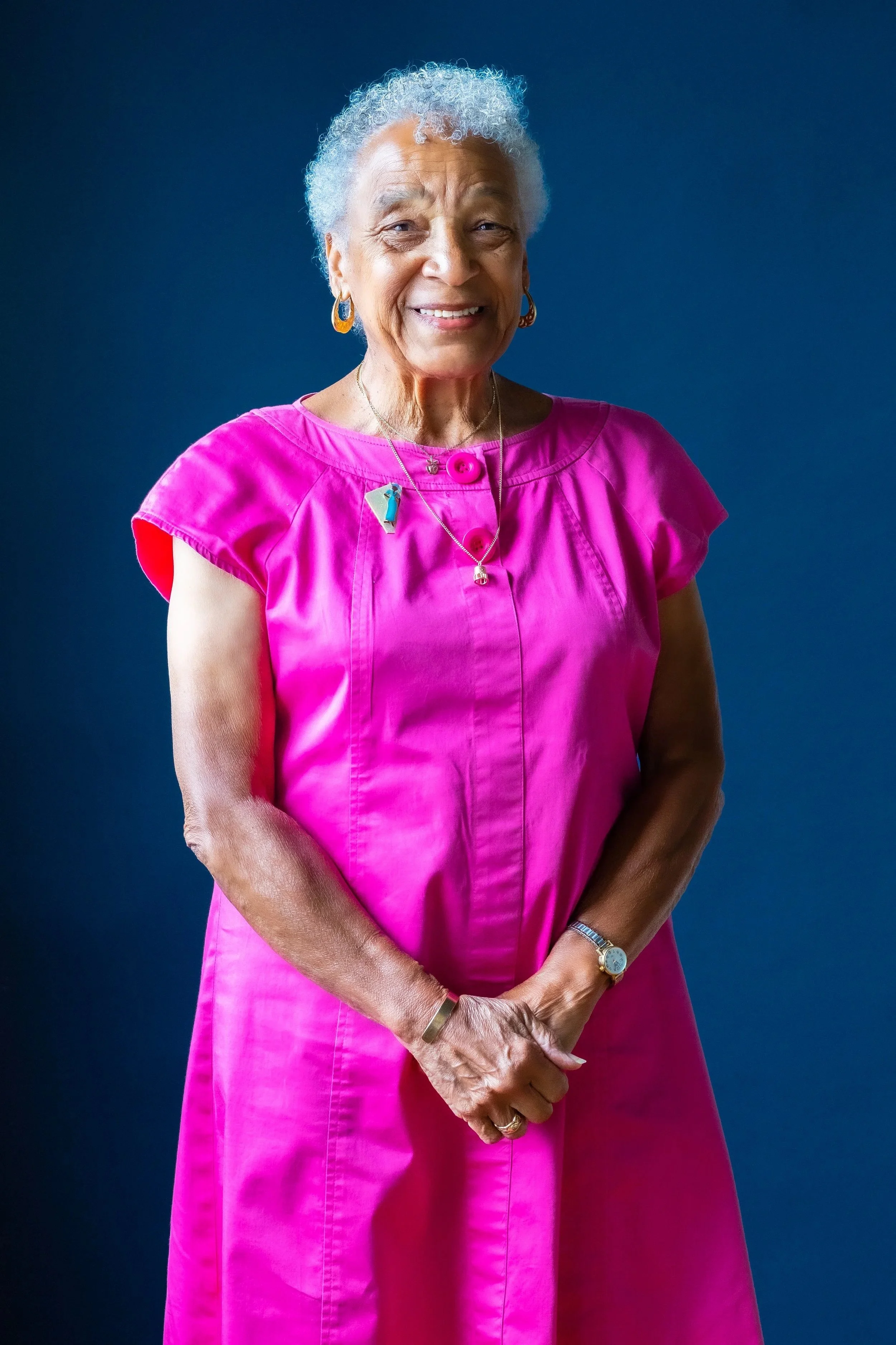 An elderly woman with gray hair tied back, smiling, wearing a bright pink dress with gold earrings, a necklace, a watch, and a bracelet, standing against a dark blue background.