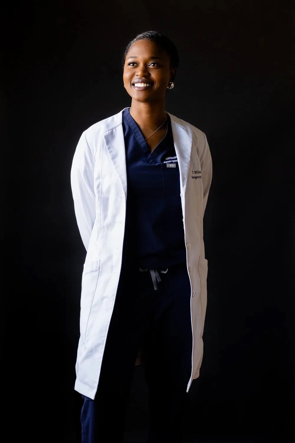 A smiling female healthcare professional standing against a dark background, wearing a white lab coat over navy scrubs.
