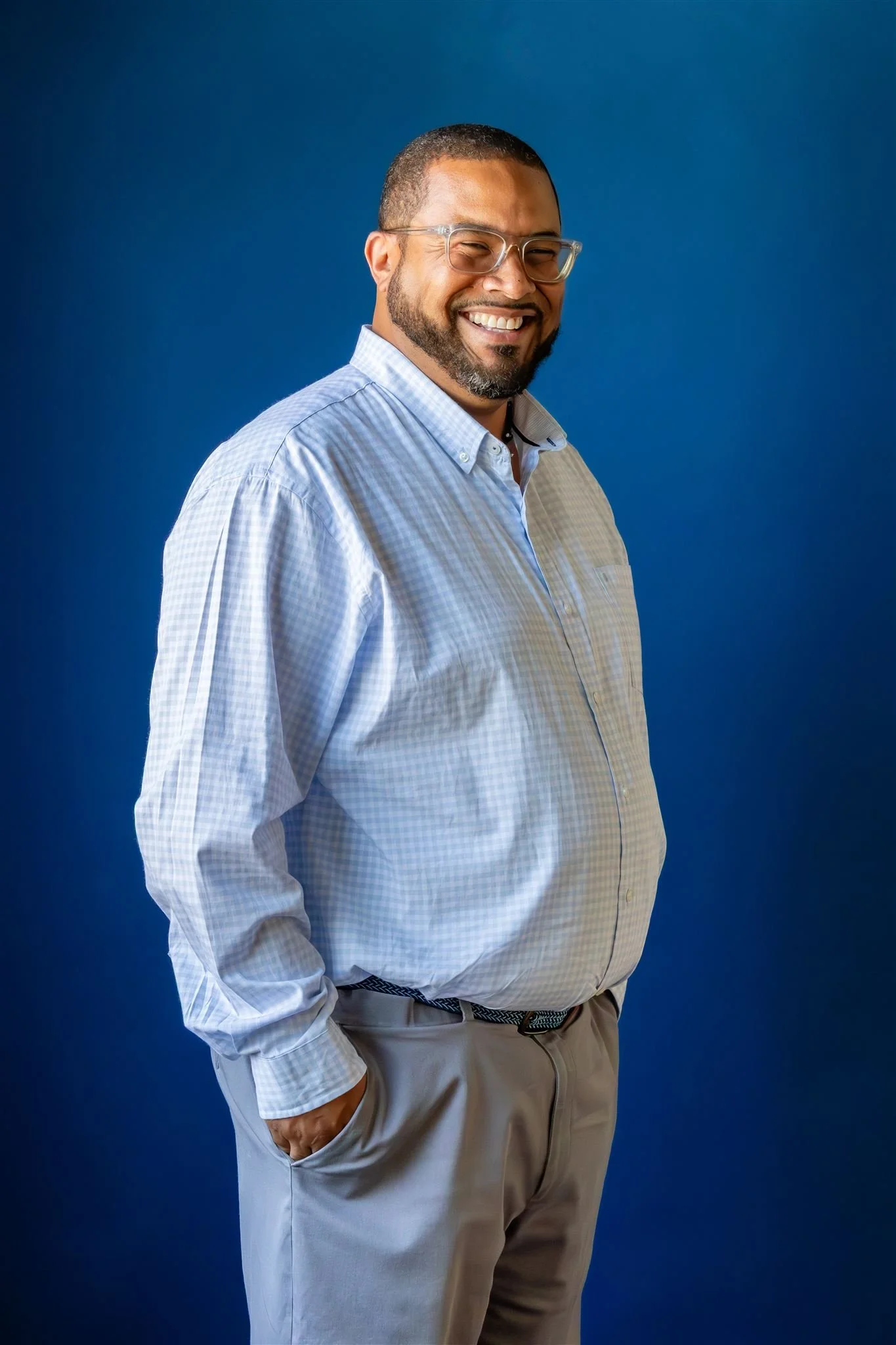 A smiling man with glasses, a beard, and short hair, wearing a light blue checked shirt and beige pants, standing against a plain blue background.