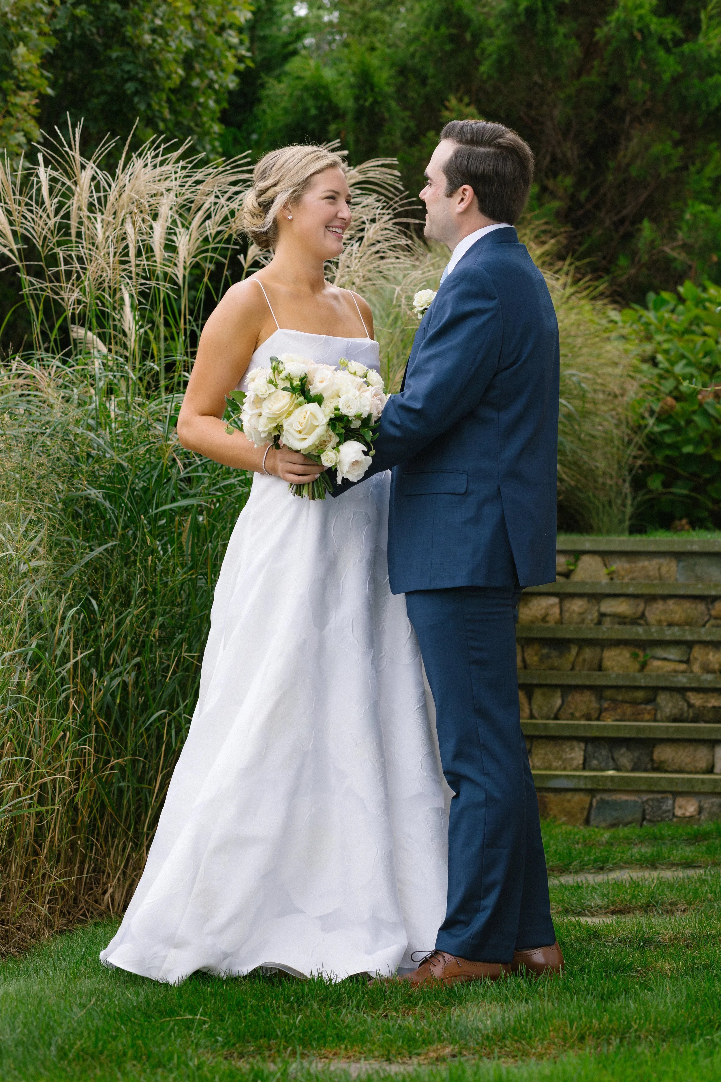 A bride and groom standing outdoors, holding hands and gazing at each other, surrounded by greenery and tall grasses, during their wedding ceremony.