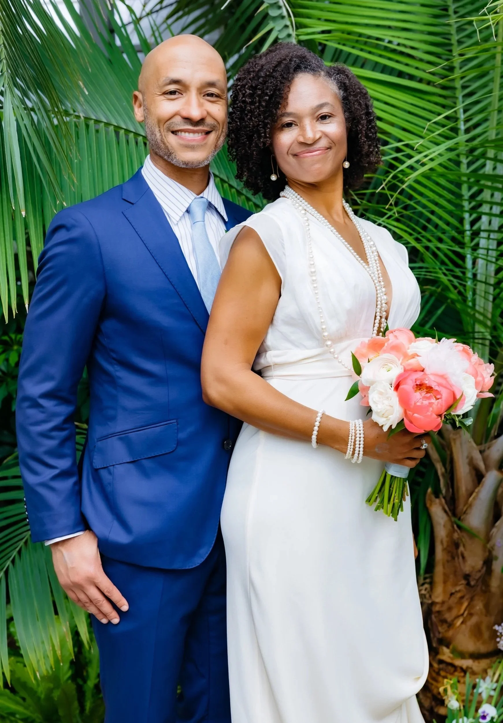 A smiling couple dressed in formal attire, the man in a blue suit and the woman in a white dress, standing in front of lush green tropical foliage. The woman holds a bouquet of pink and white flowers and wears pearl jewelry.