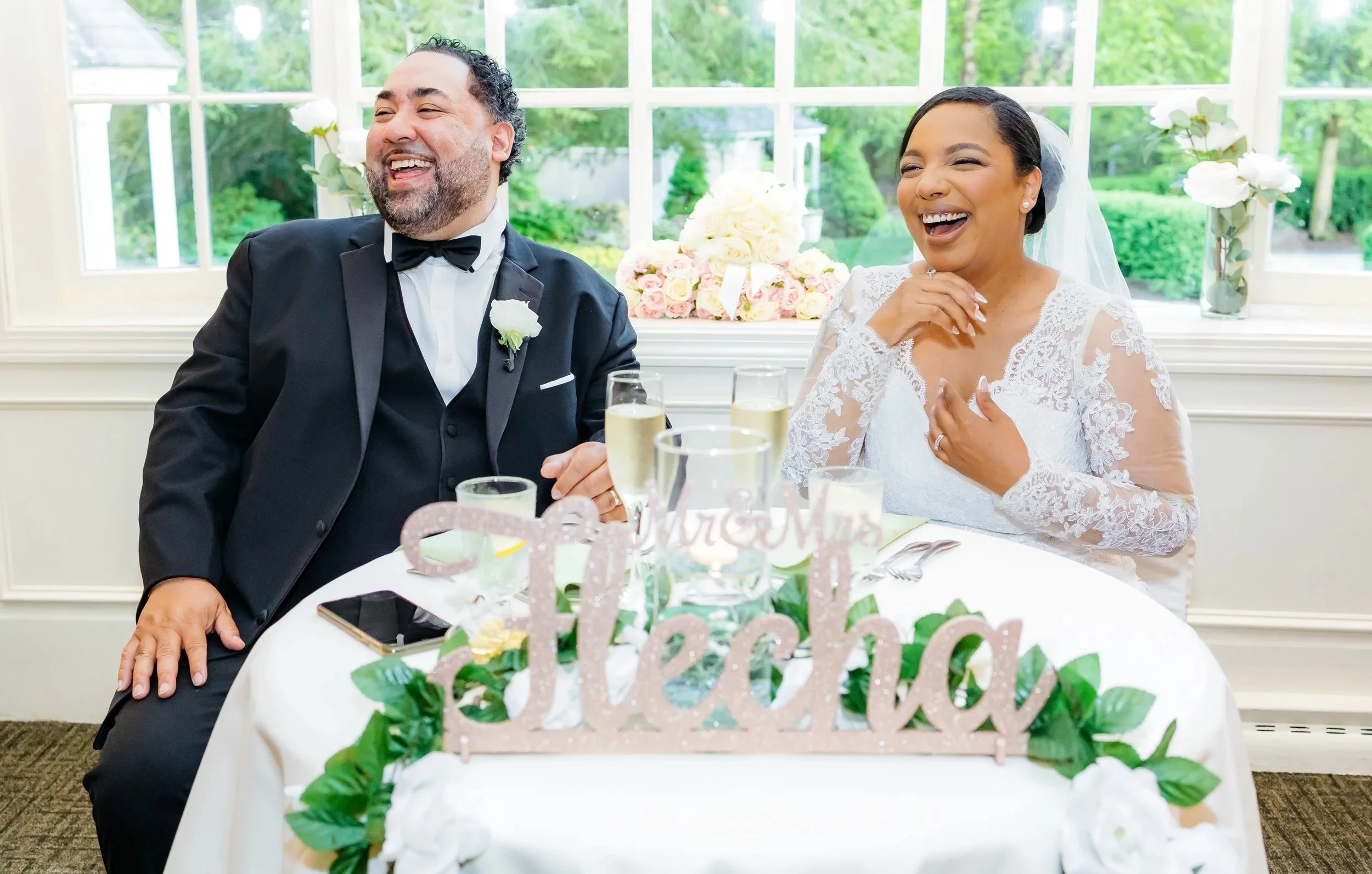 A bride and groom sit at a table, laughing and smiling during their wedding celebration. The table is decorated with green leaves and a pink "Hecho" sign, with a window and greenery outside in the background.