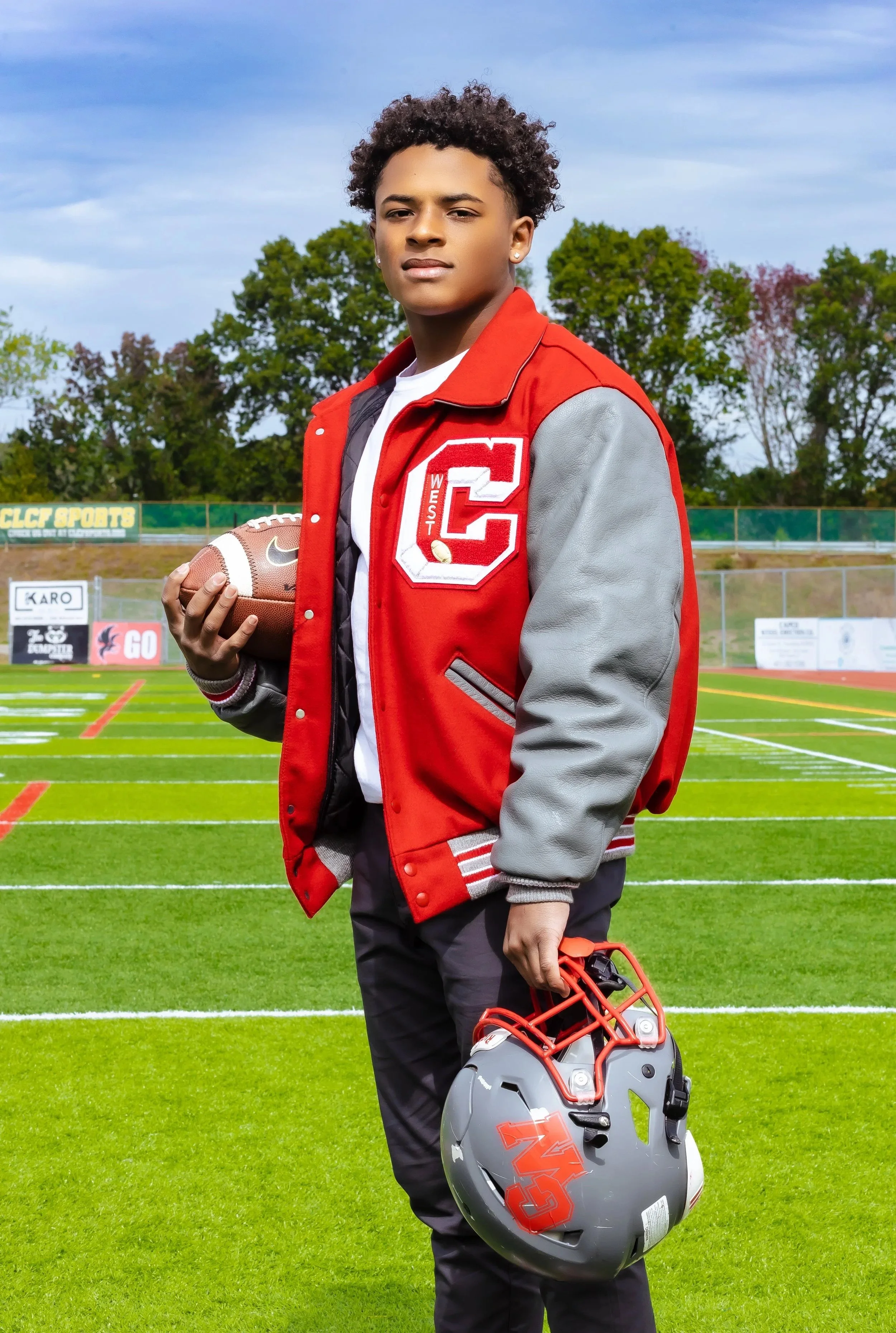 Young male football player standing on a football field, holding a football in one hand and a gray helmet with a red faceguard in the other. He is wearing a red and gray letterman jacket with a large letter 'C' on the front, and has a serious express