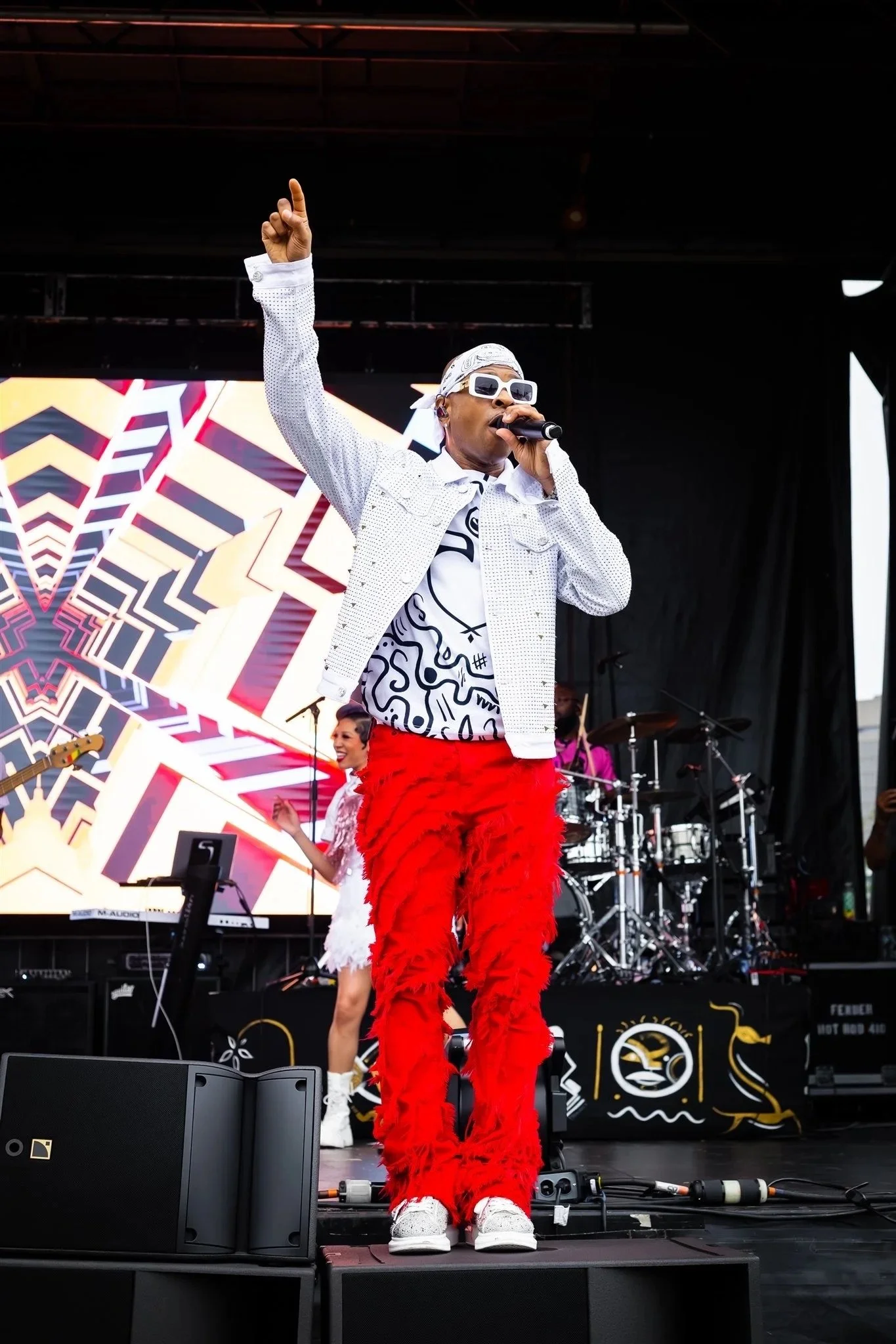 A performer on stage wearing a white bandana, white sunglasses, a white jacket with black dots, a graphic black and white shirt, and red feathered pants, holding a microphone, with a band playing in the background and a colorful digital screen behind