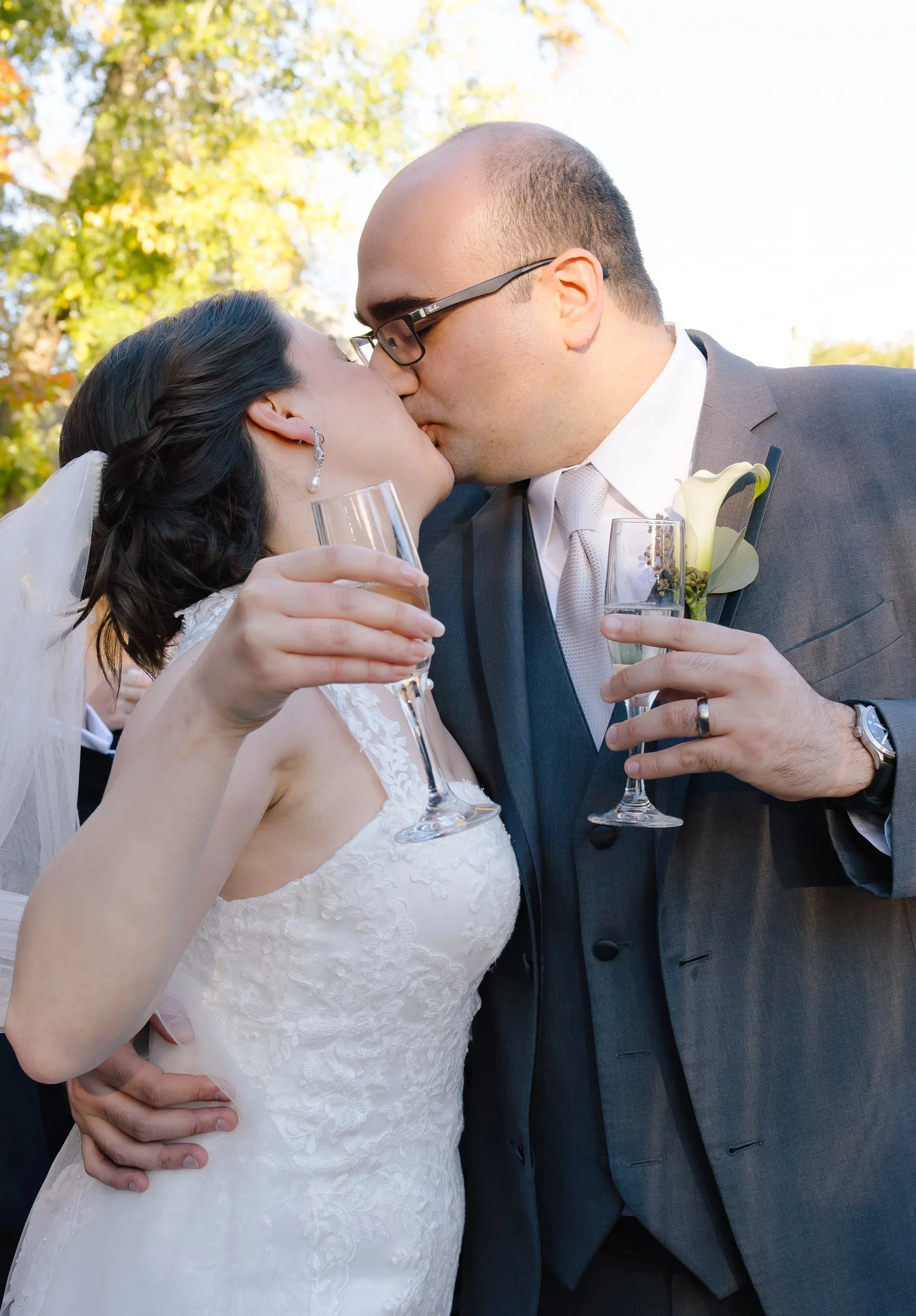 A newly married couple sharing a kiss at their outdoor wedding, holding champagne glasses.